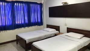 Une chambre avec deux lits simples, un grand téléviseur à écran plat fixé au mur et un climatiseur placé au-dessus de la fenêtre, le tout agrémenté de rideaux bleus.