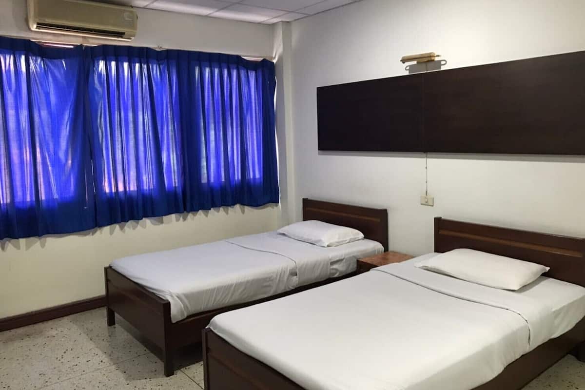 A room with two single beds, a large dark TV mounted on the wall, and an air conditioner above the window with blue curtains.
