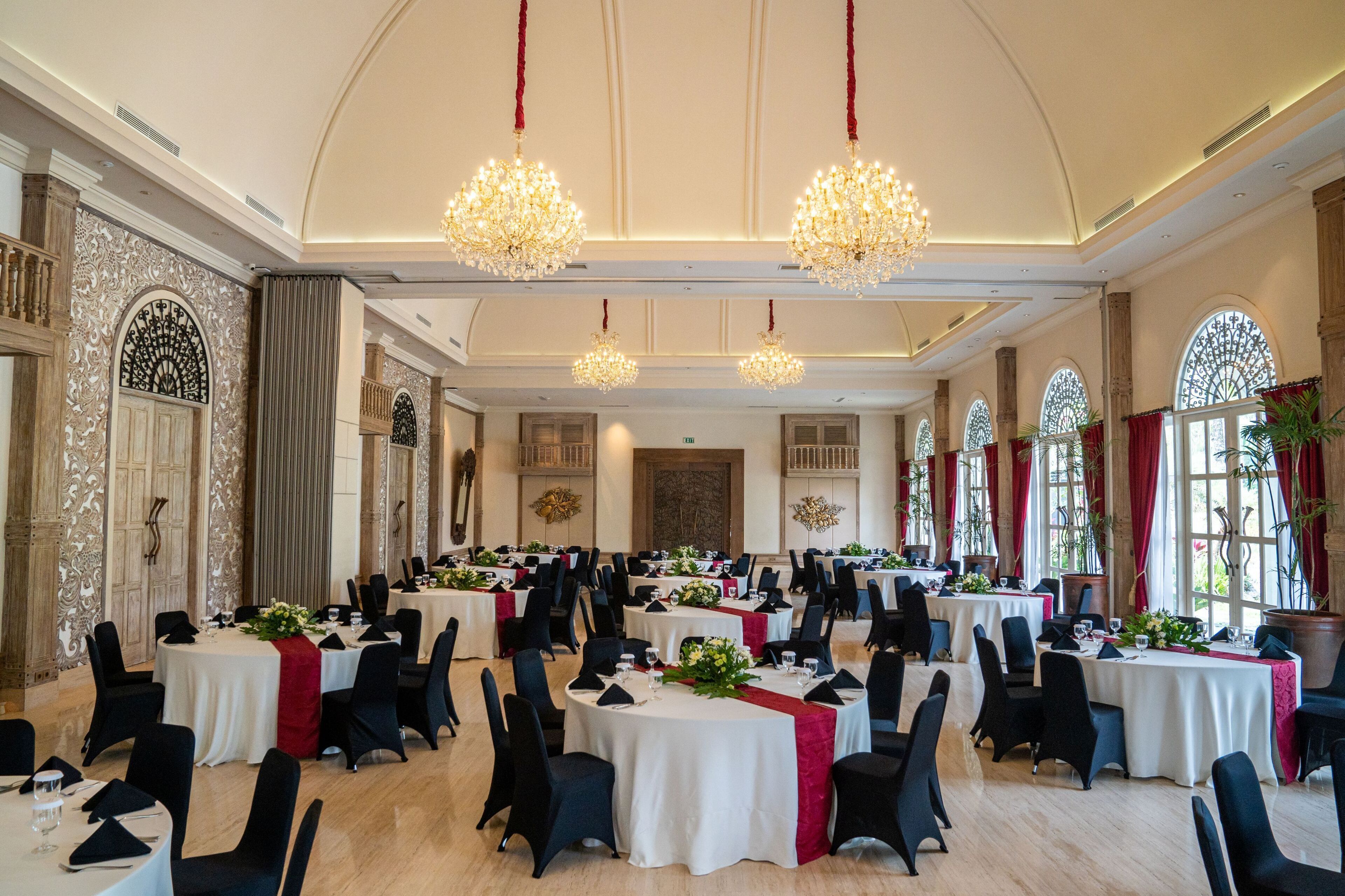 A banquet hall with round tables, black chairs, chandeliers, and large windows with red curtains.