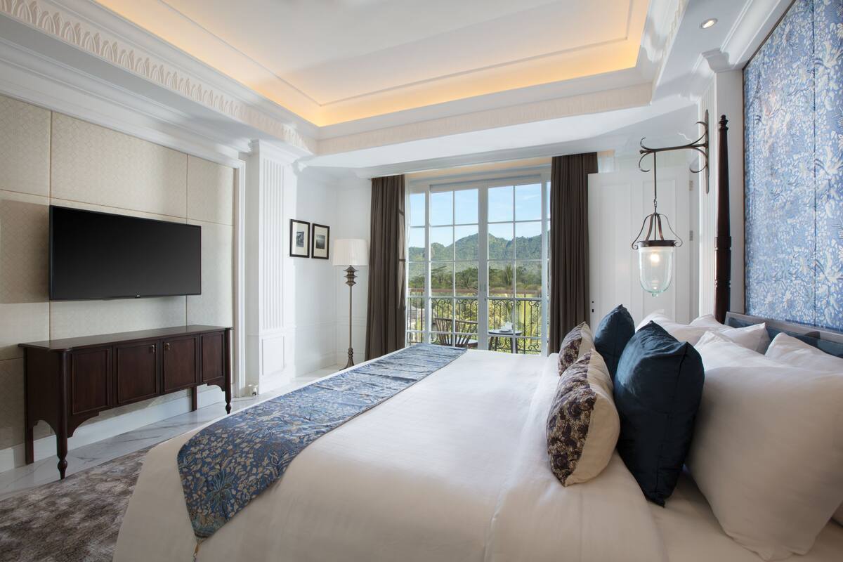 A spacious bedroom with a large bed, a flat-screen TV mounted on the wall, a balcony with a view of the mountains, and a chandelier.