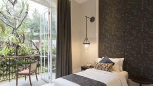 A hotel room with a bed, a nightstand, a chair, and a balcony with plants. - Plataran Heritage Borobudur Hotel (Borobudur)