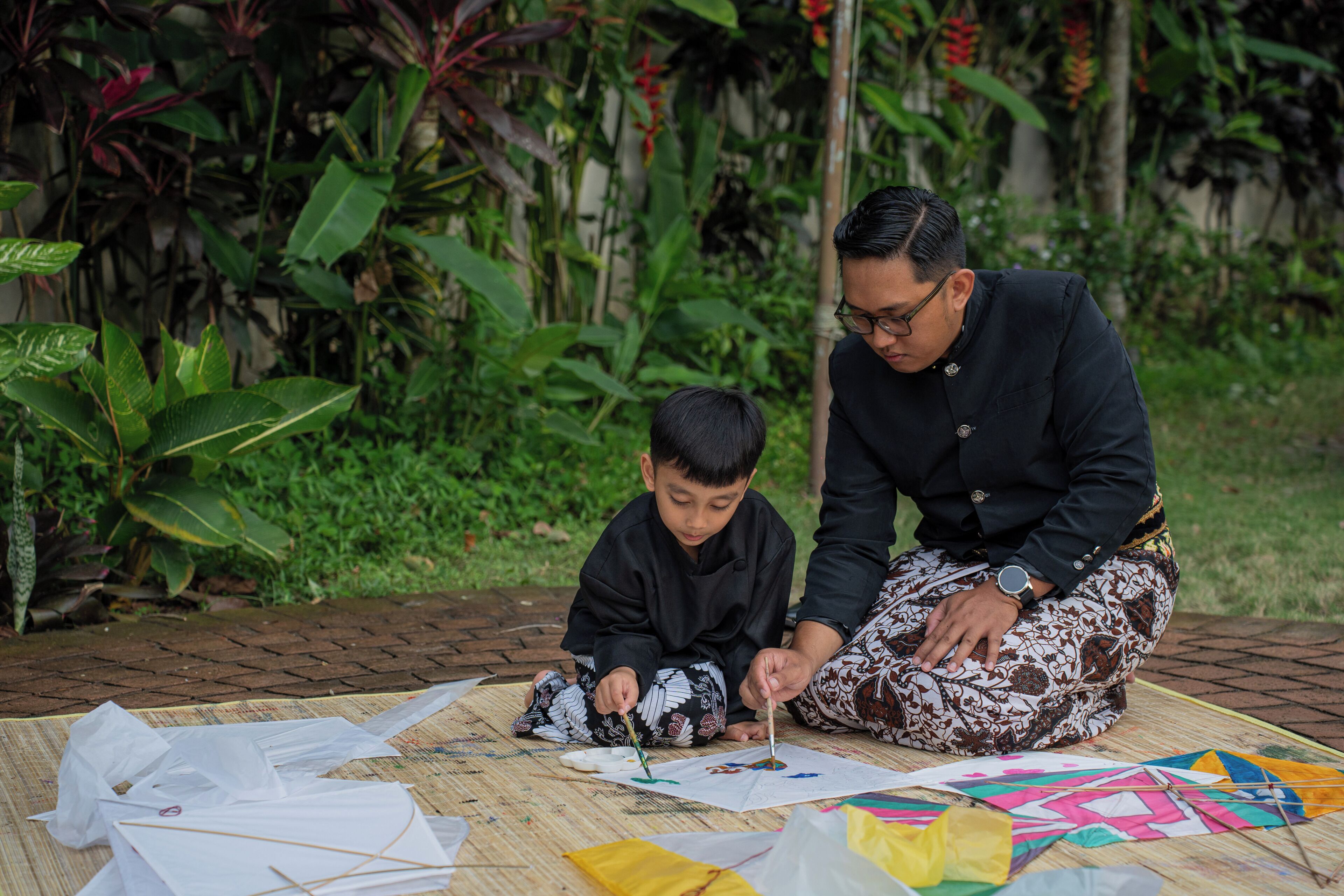Two individuals sitting on a mat outdoors with various papers and kites, surrounded by greenery.
