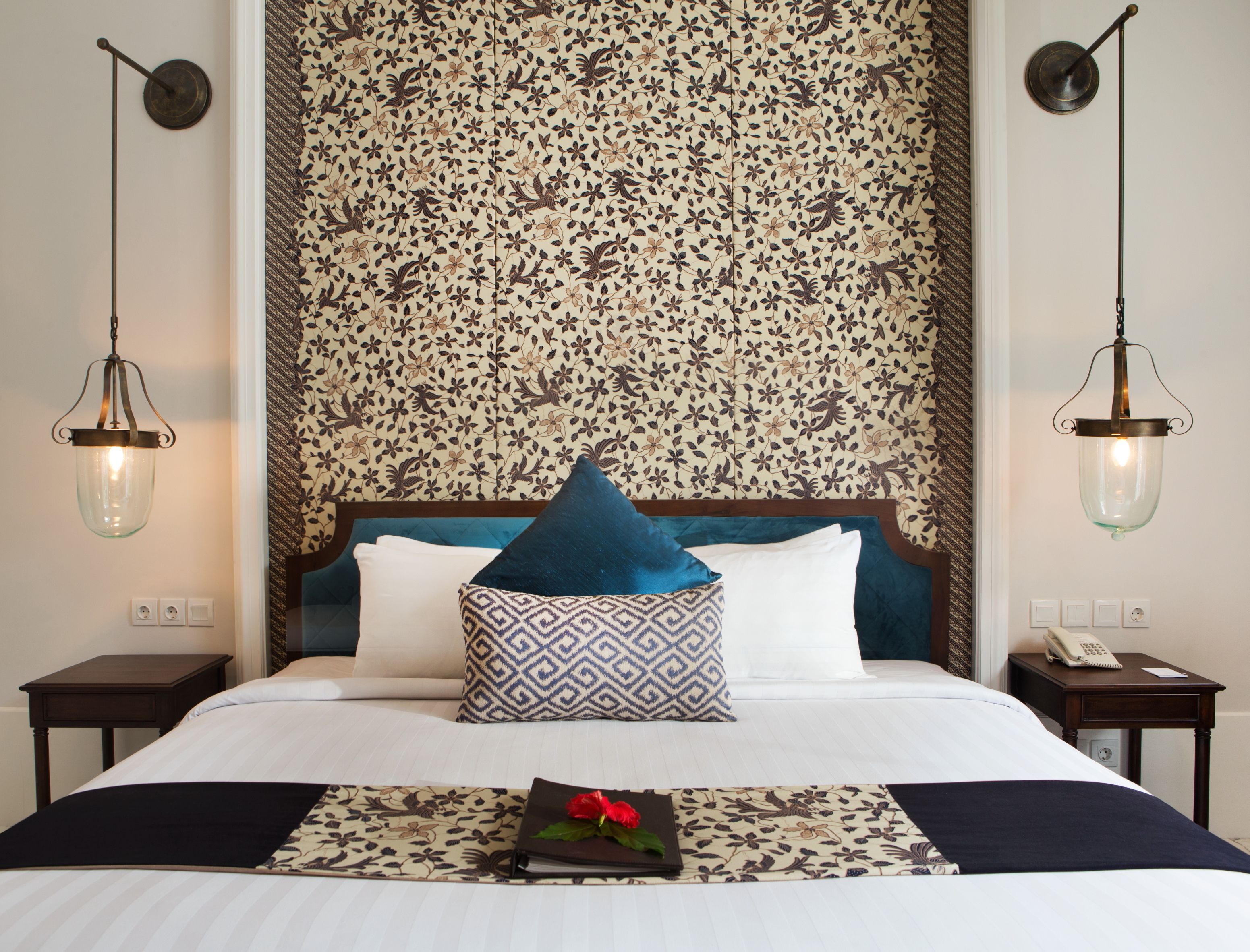 A neatly made bed with a patterned headboard, two bedside tables, and a floral wallpaper.