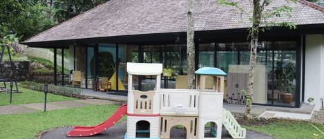 A children's play area with a slide and climbing structures, surrounded by a well-maintained lawn and a building with large windows in the background.
