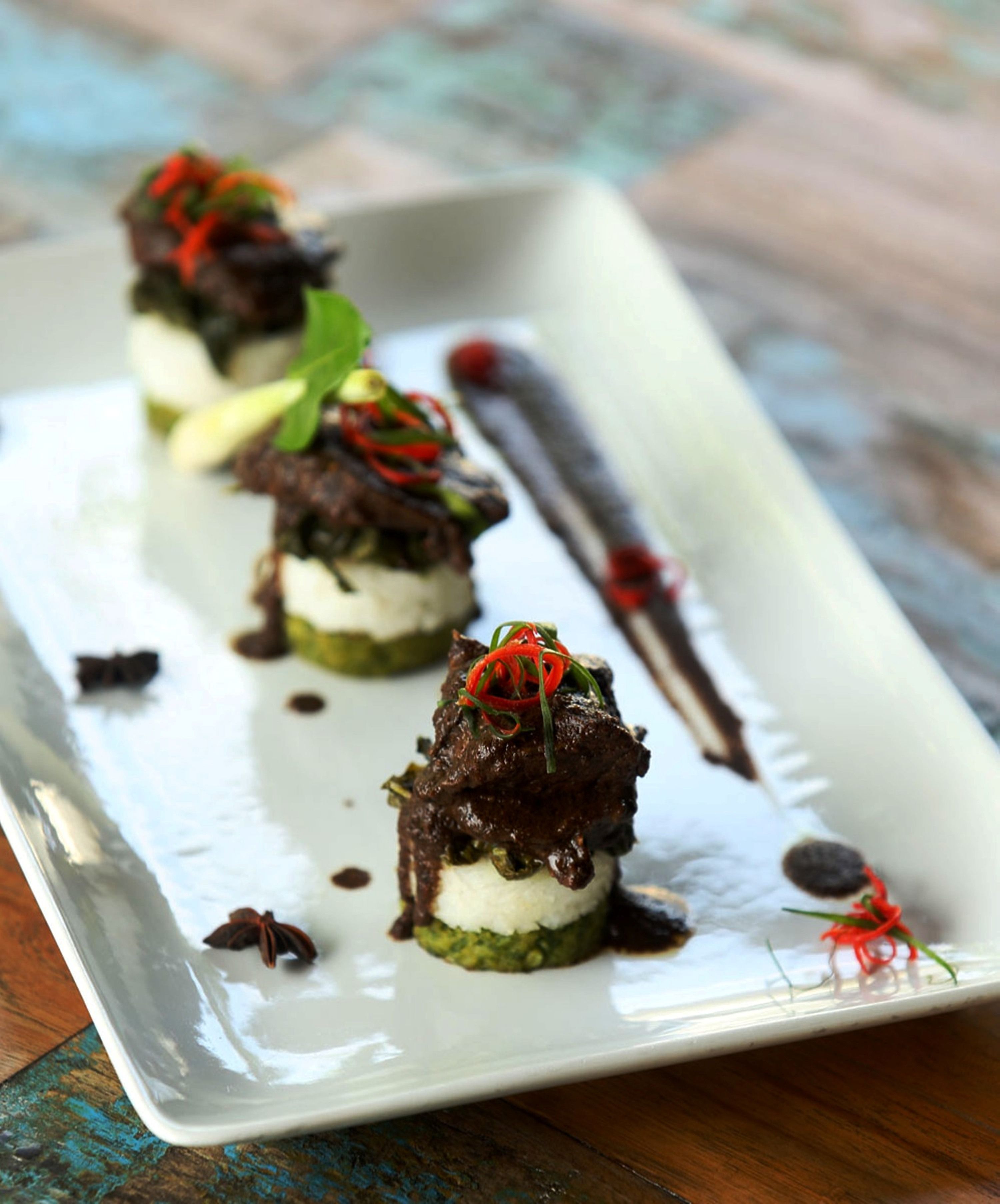 A white rectangular plate with three food items, each topped with a dark sauce and garnished with red chili and green herbs, placed on a rustic wooden table.