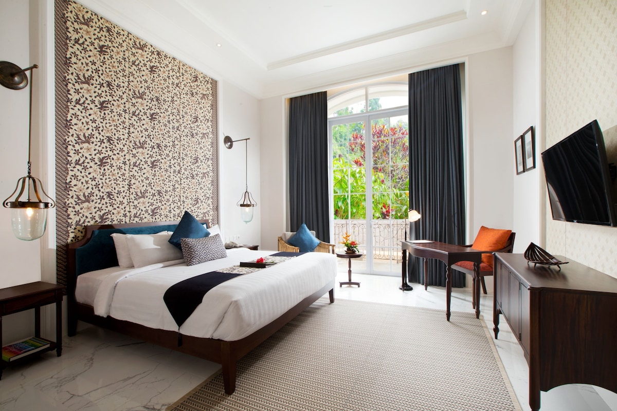 A modern hotel room with a large bed, a desk, a chair, and a balcony with a view of greenery.