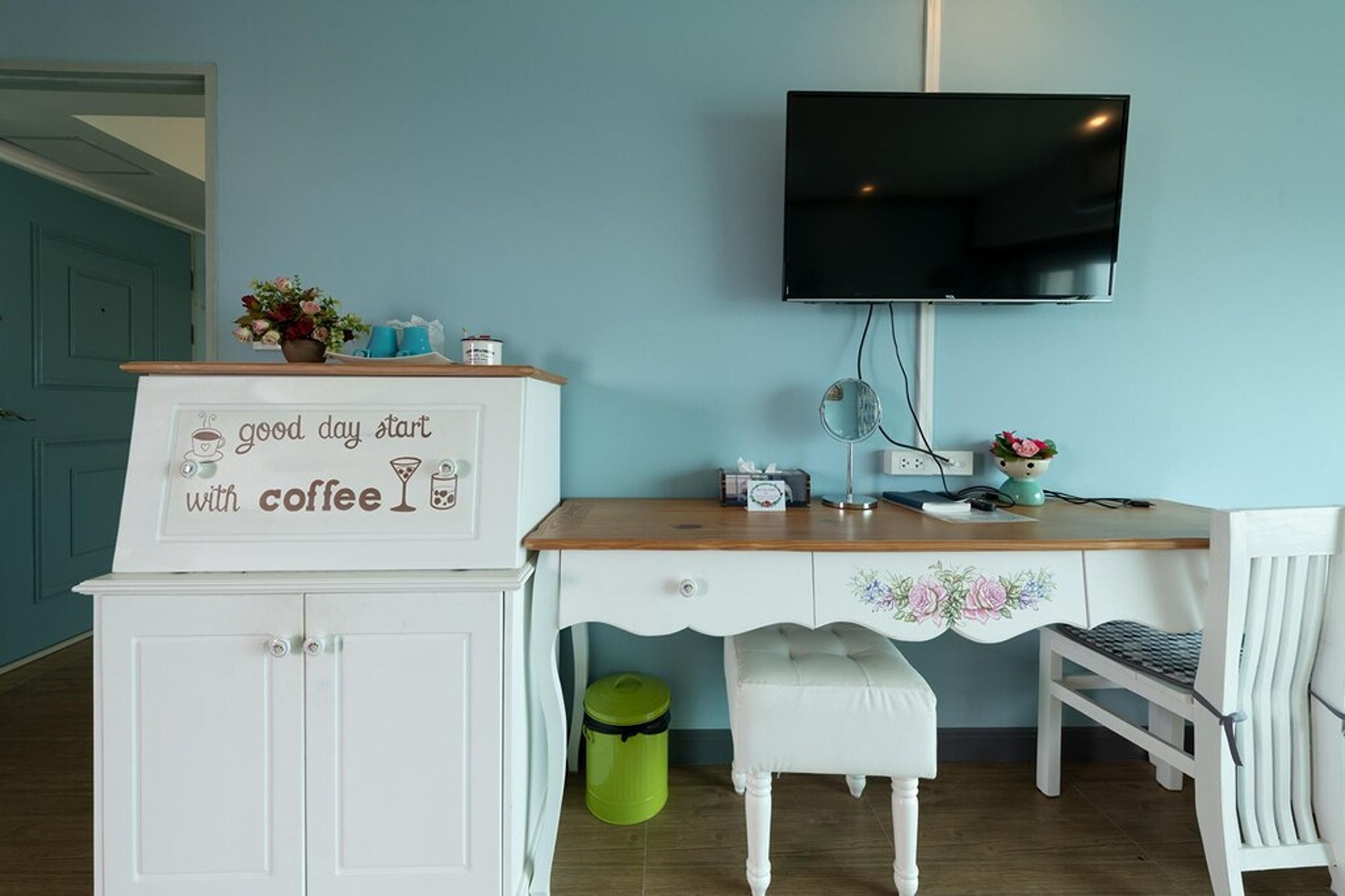 A room with a wooden desk, a white chair, a television mounted on the wall, and a cabinet with a sign saying "good day start with coffee".