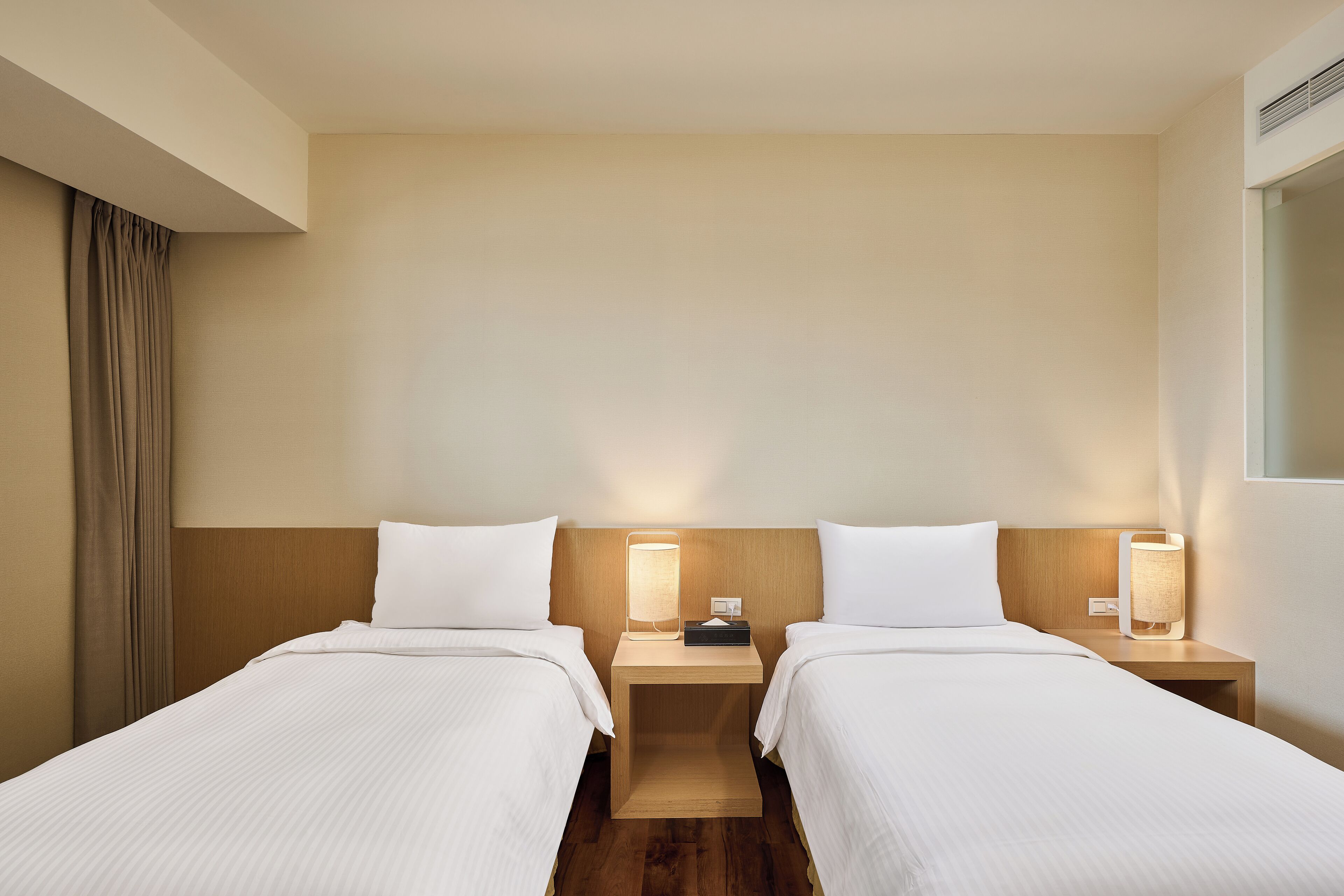Quarto de hotel com duas camas, cabeceiras de madeira, mesas de cabeceira com abajures e uma paleta de cores neutras.
