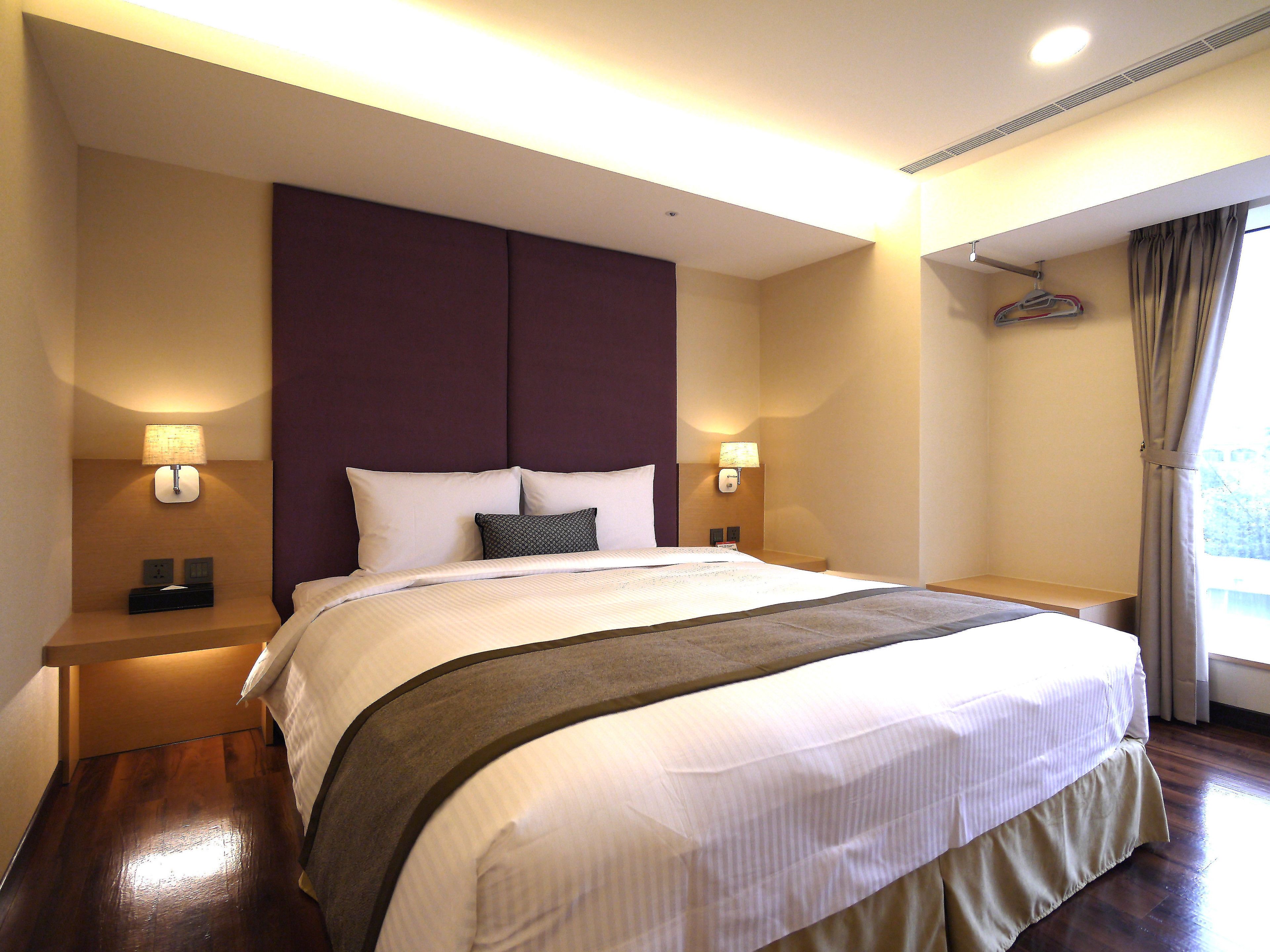 A hotel room with a large bed, two bedside tables, a wall-mounted lamp, and a window with curtains.