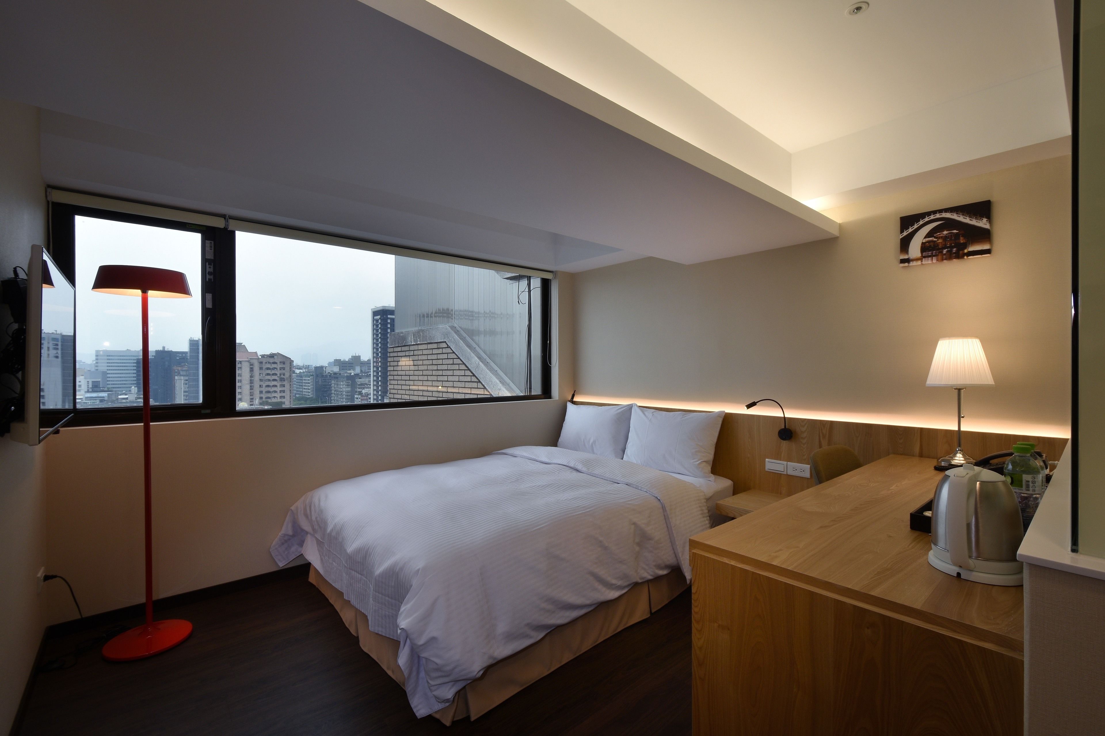 A modern hotel room with a bed, a desk, a red floor lamp, a white lamp, and a city view through large windows.