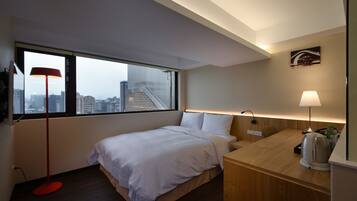 A modern hotel room with a bed, a desk, a red floor lamp, a white lamp, and a city view through large windows.