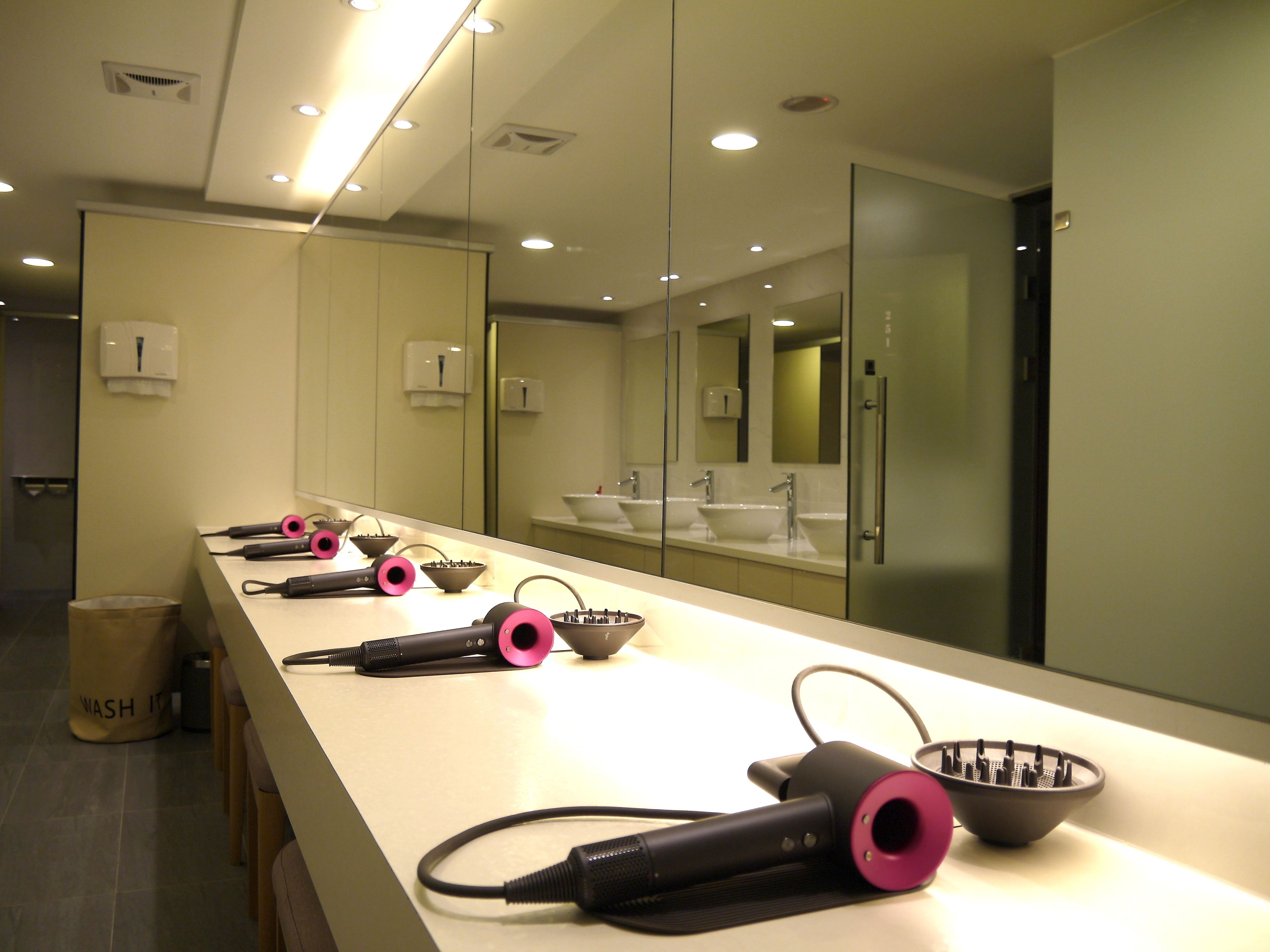 A modern bathroom with a long counter, multiple sinks, and hair dryers.