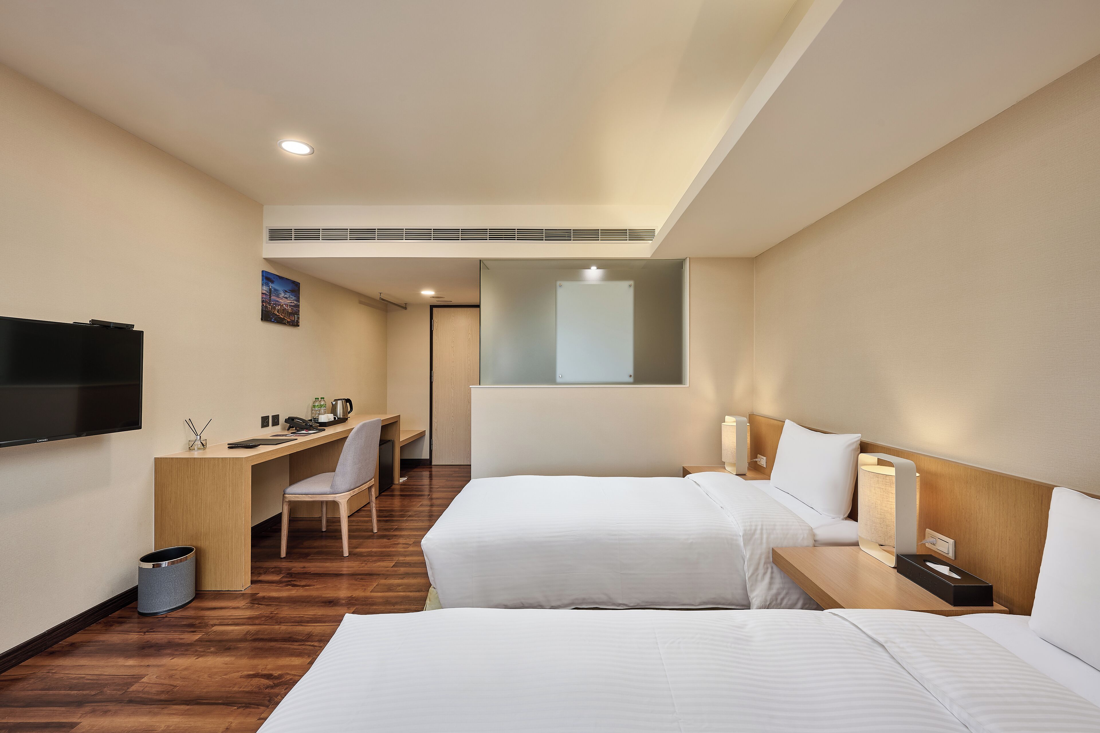 A hotel room with two beds, a desk, a chair, a TV, and a window.