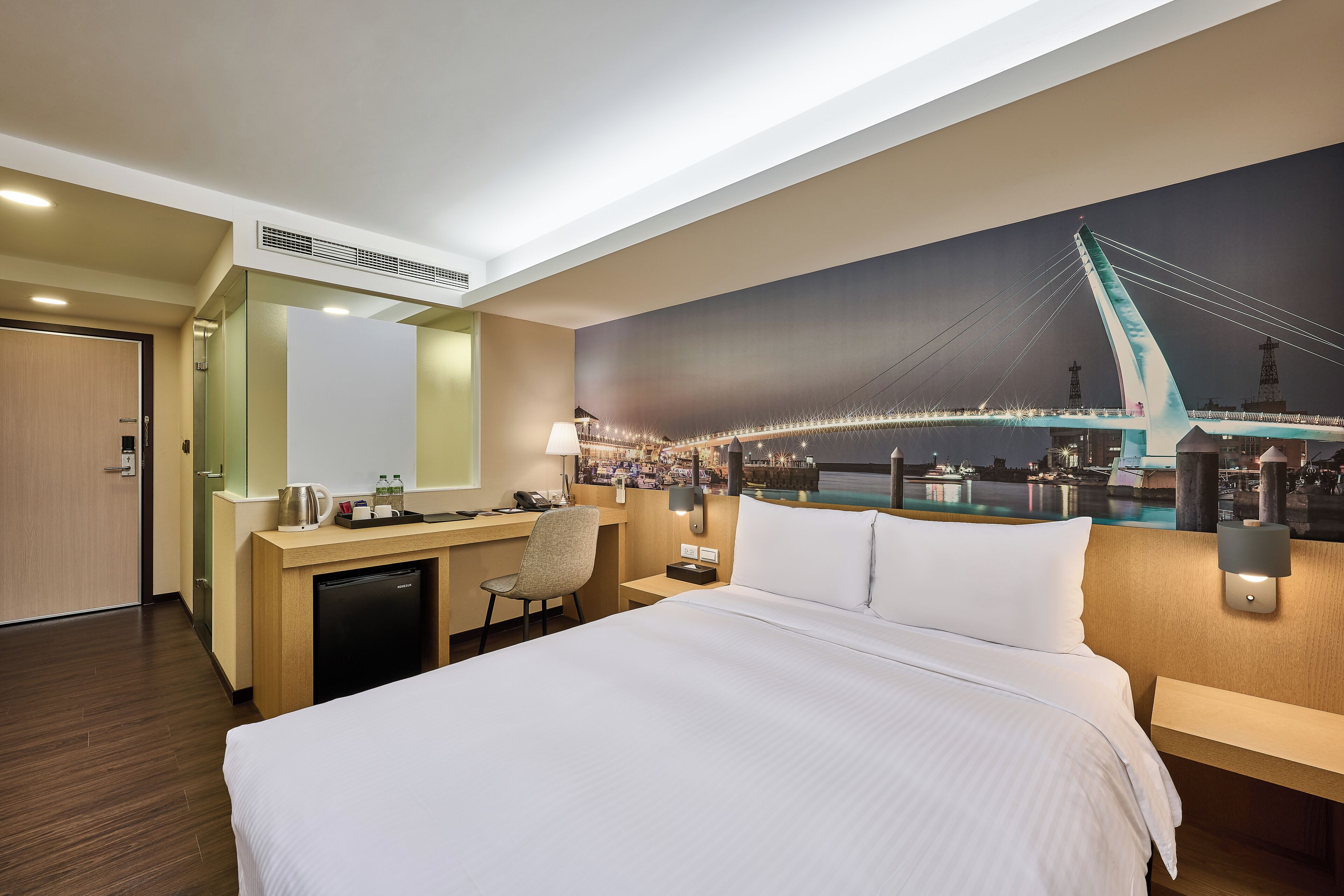 A hotel room with a bed, desk, chair, and a large window with a cityscape view.