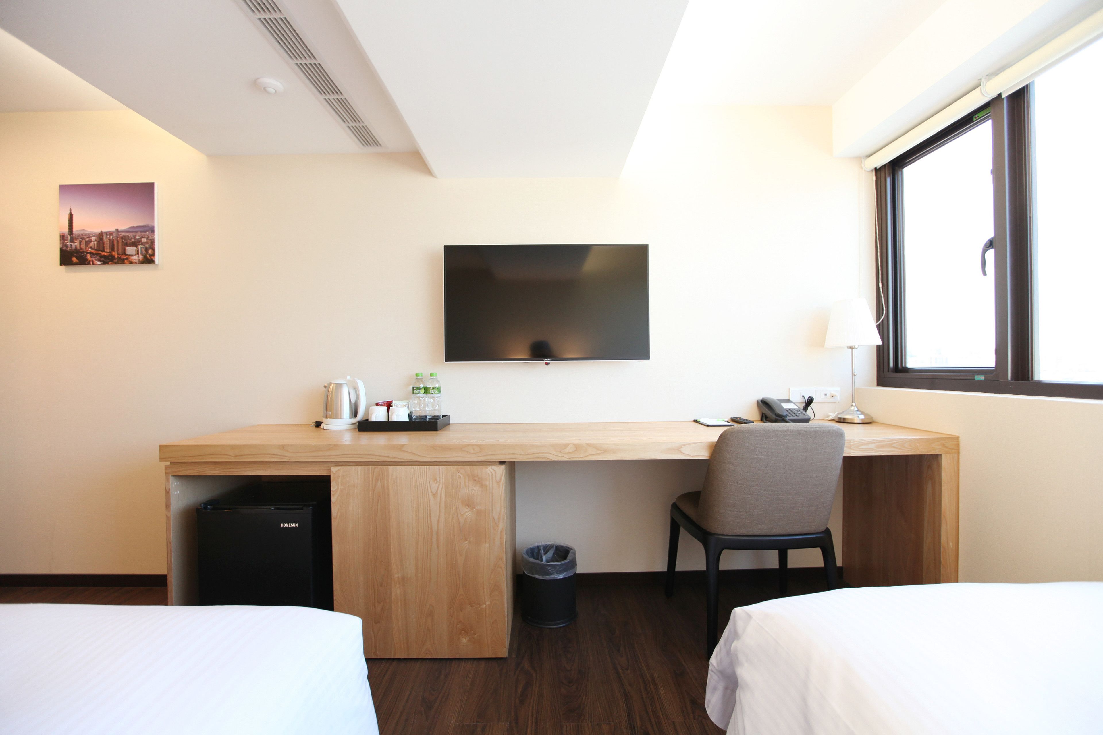 A hotel room with a wooden desk, a chair, a bed, a TV, and a window.