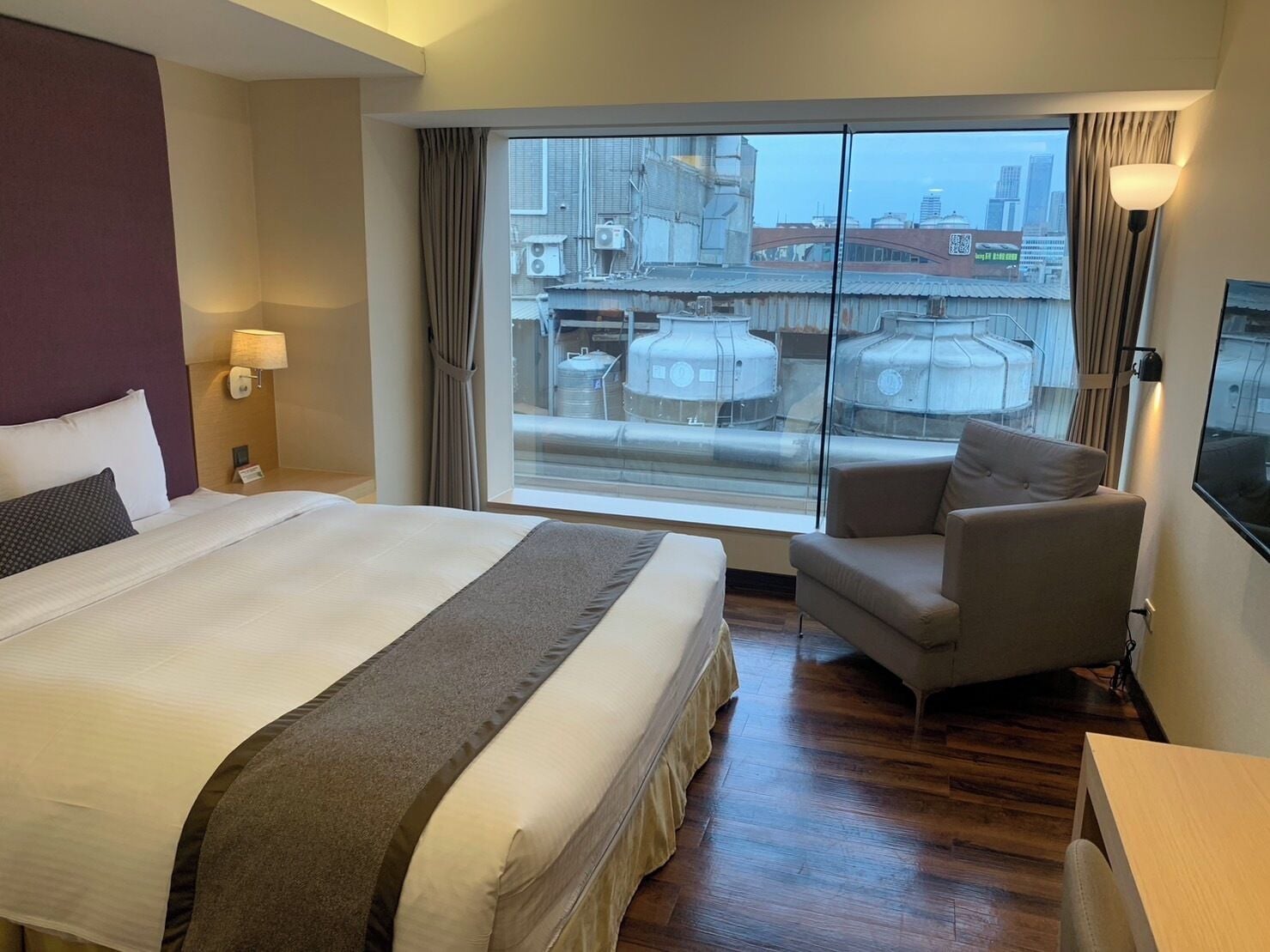 A hotel room with a large bed, a chair, a desk, and a view of the city through a large window.