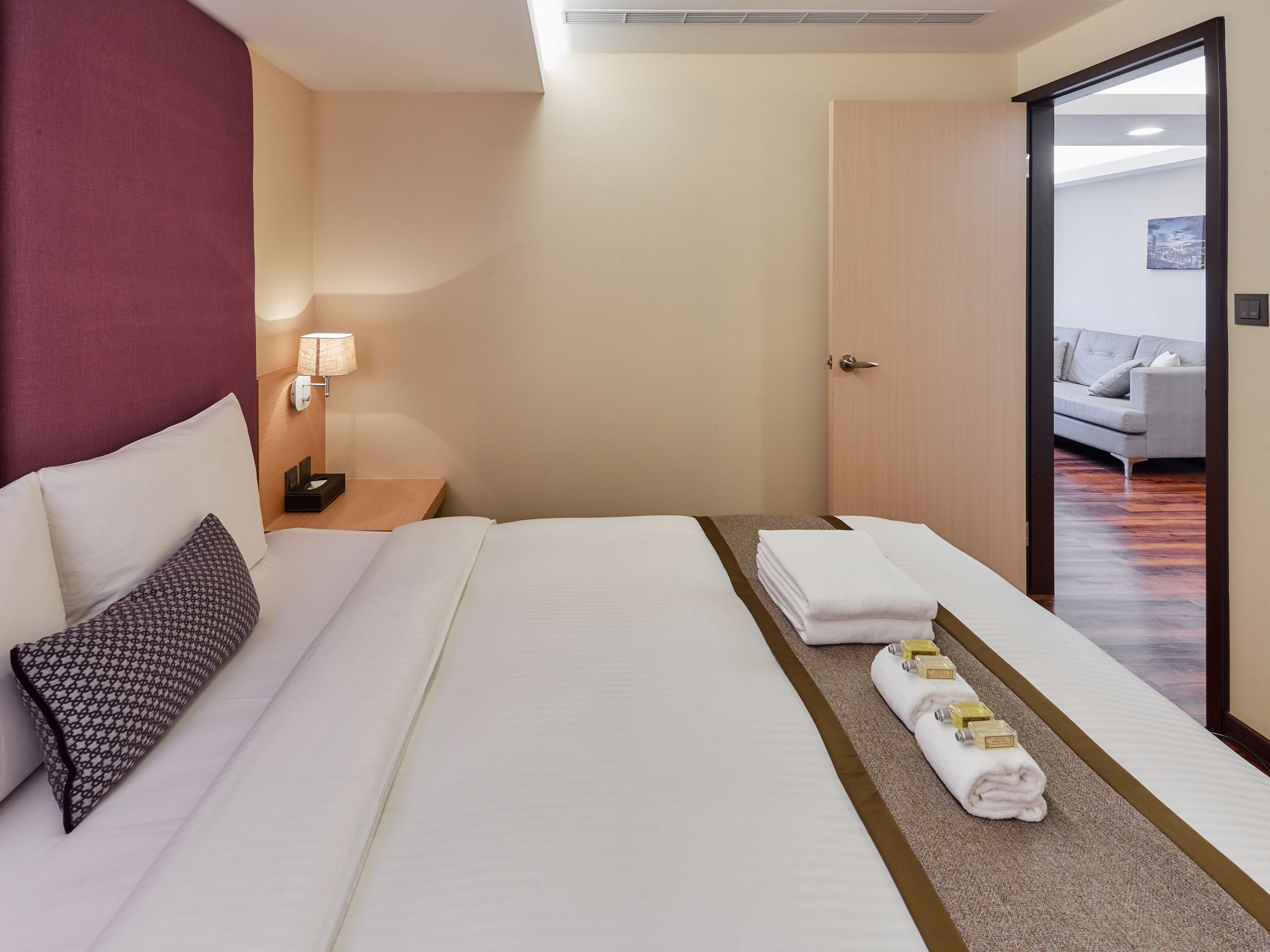 A hotel room with a large bed, a nightstand, a lamp, and a view of a living area through an open door.