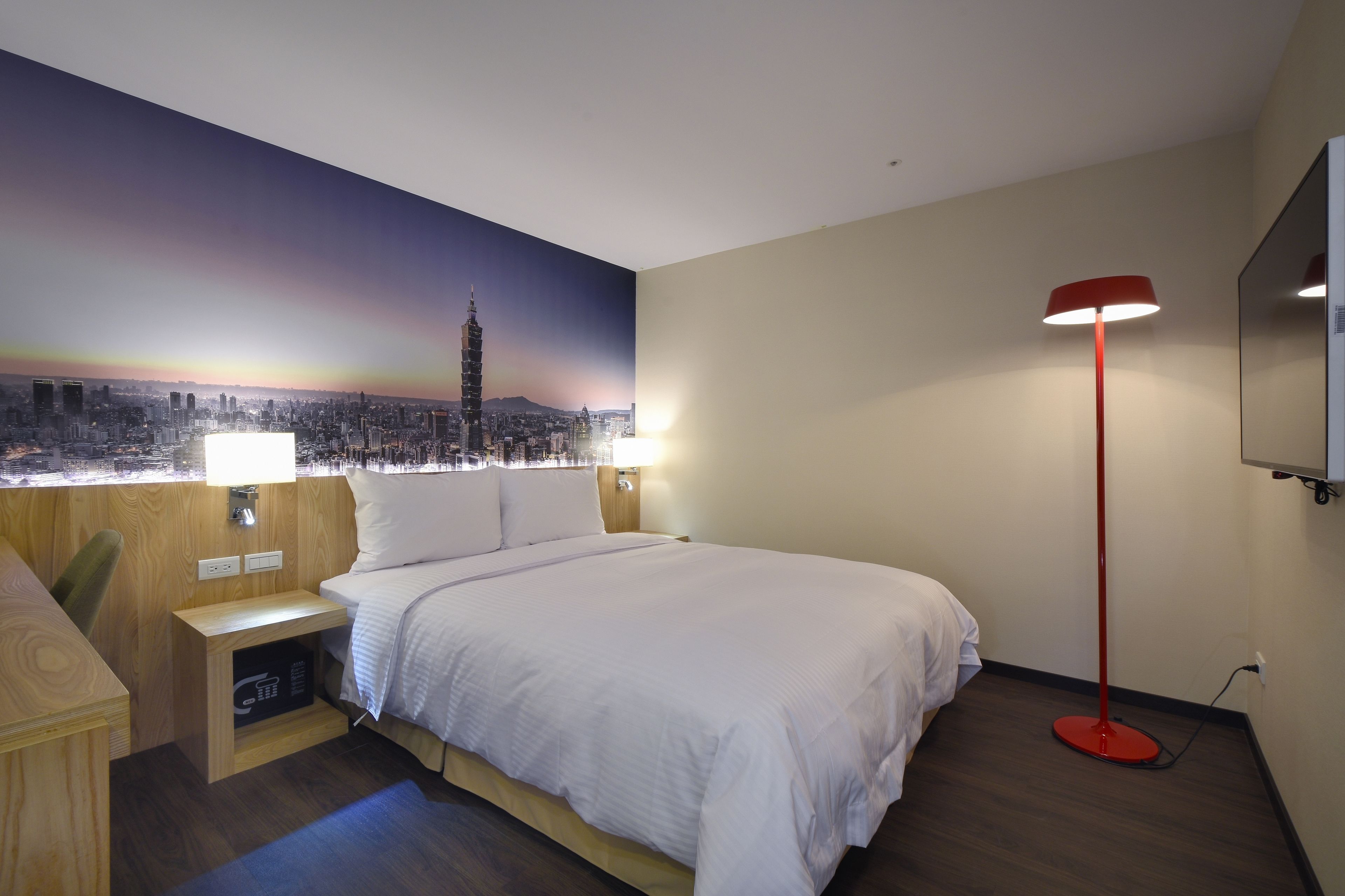 A hotel room with a bed, a desk, a red floor lamp, a TV, and a cityscape mural on the wall.