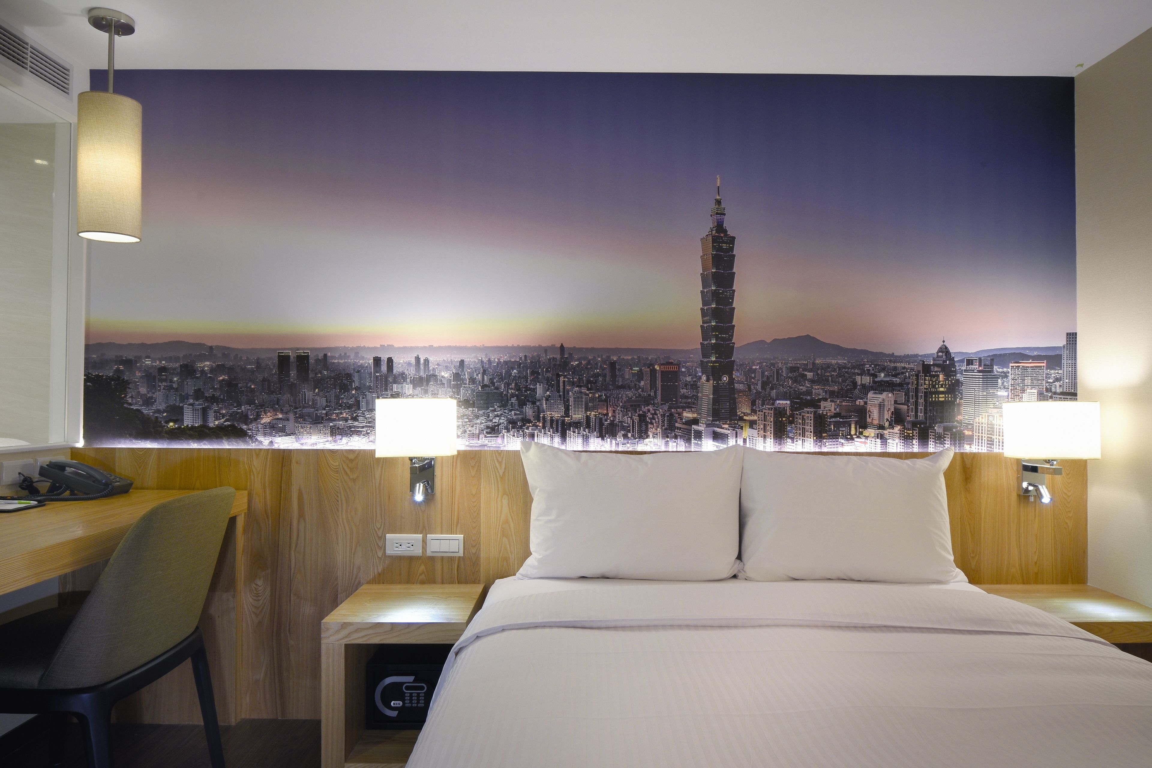A hotel room with a bed, two bedside tables, a desk, and a chair. There is a cityscape mural on the wall behind the bed.