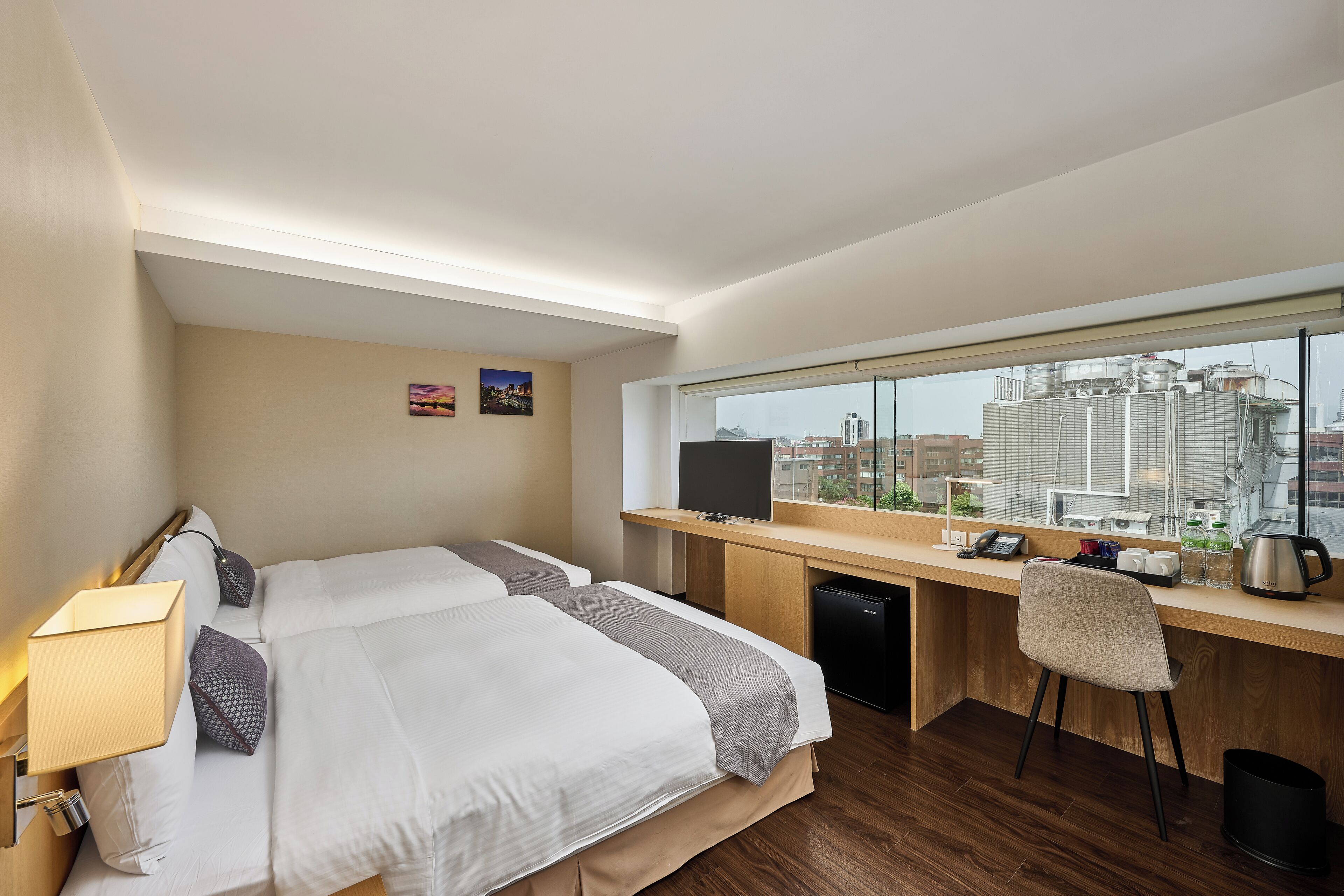 A hotel room with two beds, a desk with a computer, a chair, a TV, and a window with a city view.