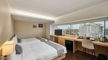 A hotel room with two beds, a desk with a computer, a chair, a TV, and a window with a city view.