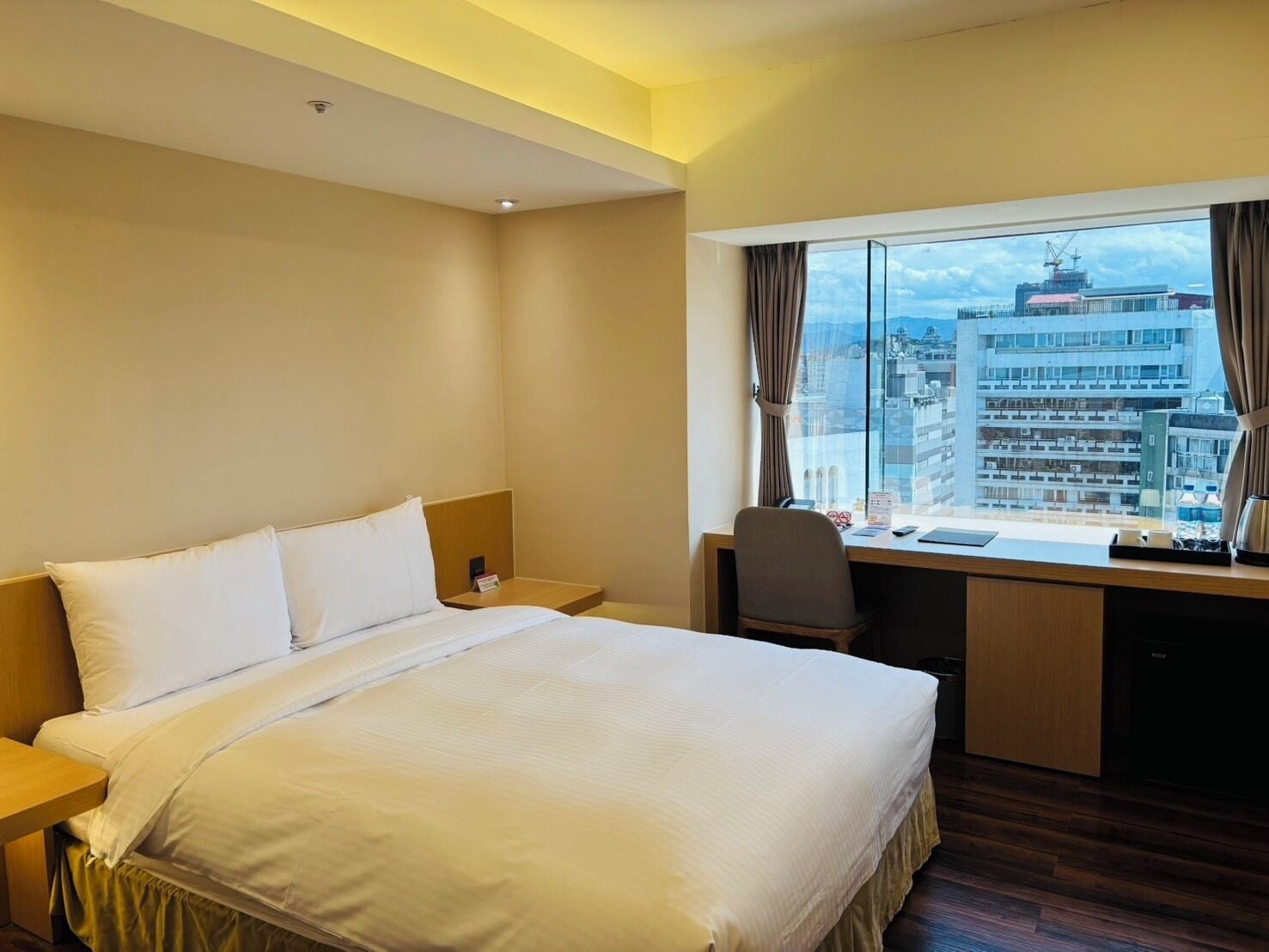 Habitación de hotel con cama, escritorio y vistas a un paisaje urbano a través de amplias ventanas.