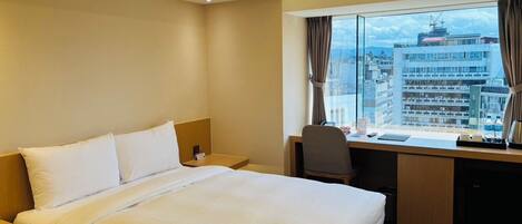 Habitación de hotel con cama, escritorio y vistas a un paisaje urbano a través de amplias ventanas.