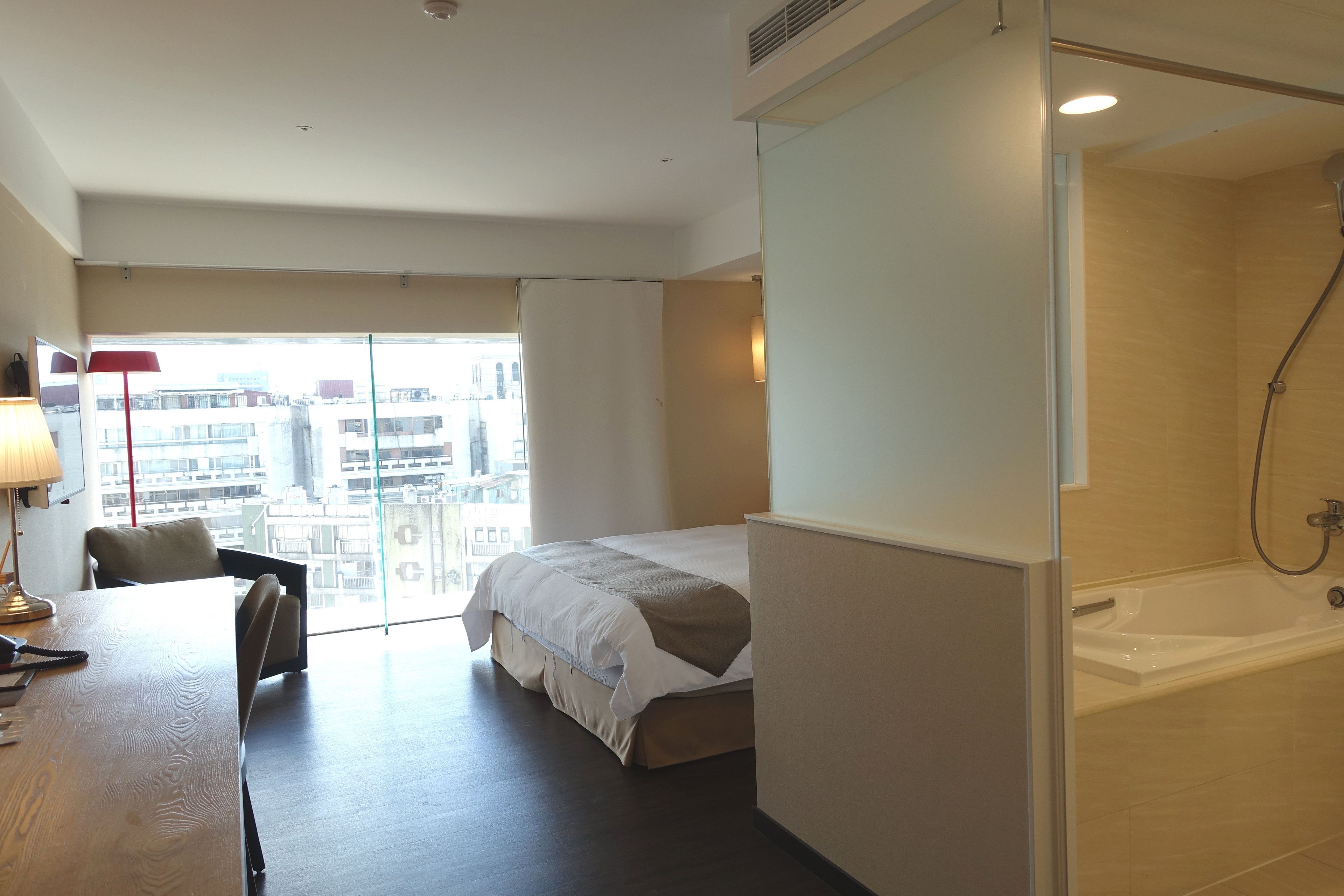 A hotel room with a bed, a desk with a lamp, a shower, and a view of buildings.