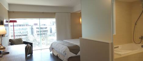 A hotel room with a bed, a desk with a lamp, a shower, and a view of buildings.