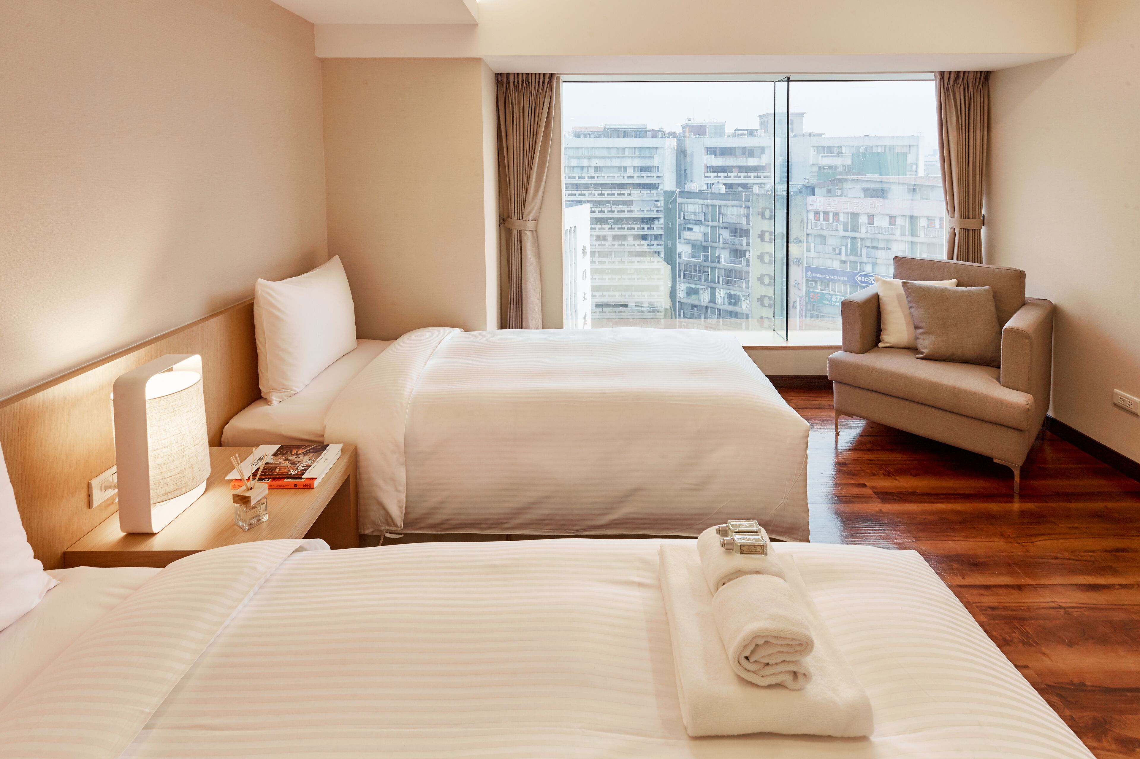 Habitación de hotel con dos camas, un sofá, una mesita de noche con lámpara y vistas a la ciudad a través de amplios ventanales.