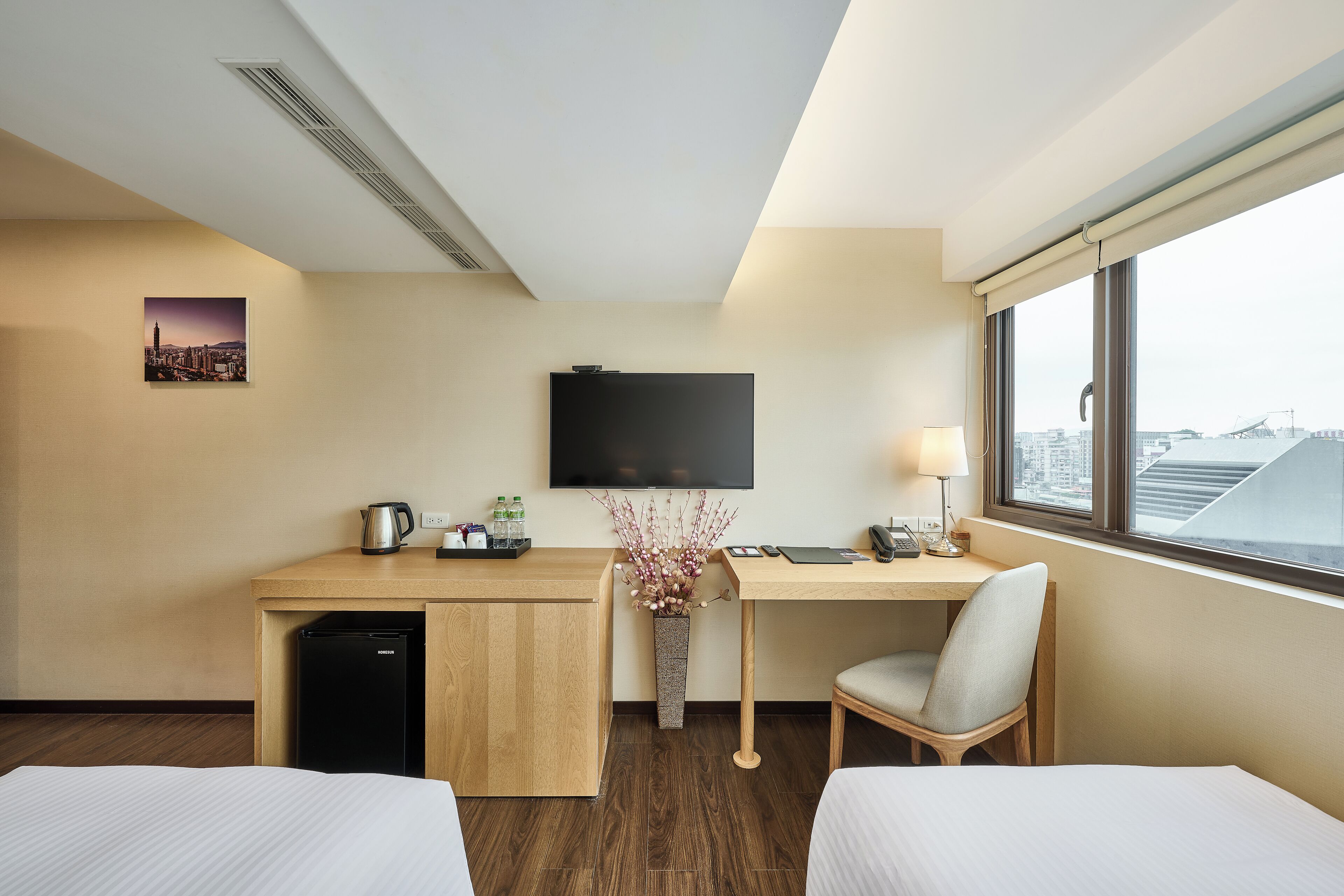 A hotel room with a bed, a desk with a computer, a TV, and a window with a city view.