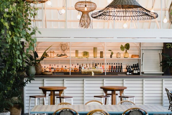 An indoor seating area with tables and chairs, a bar with shelves of bottles, and hanging light fixtures.