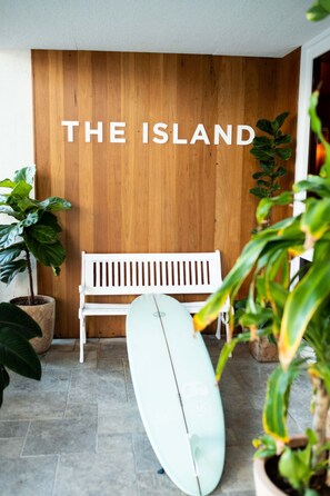 A wooden wall with "THE ISLAND" text, a white bench, and a surfboard on a tiled floor.