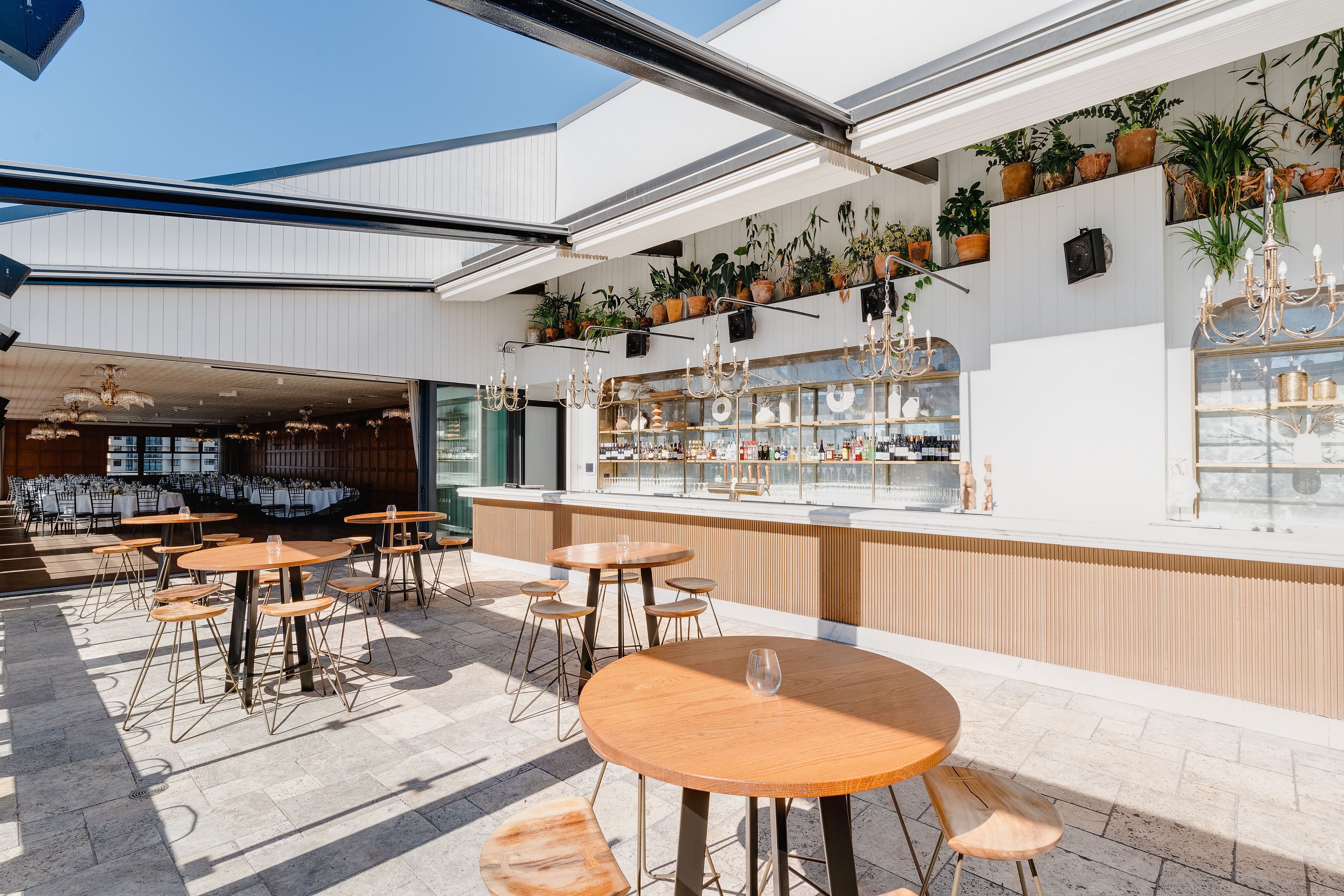 An outdoor seating area with round wooden tables and chairs, a bar with shelves of bottles, and potted plants on the wall.