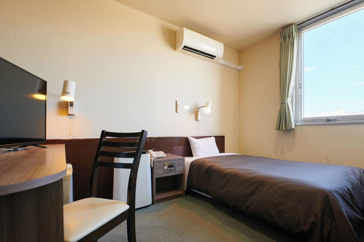 A hotel room with a bed, a desk with a chair, a TV, an air conditioner, and a window with curtains.
