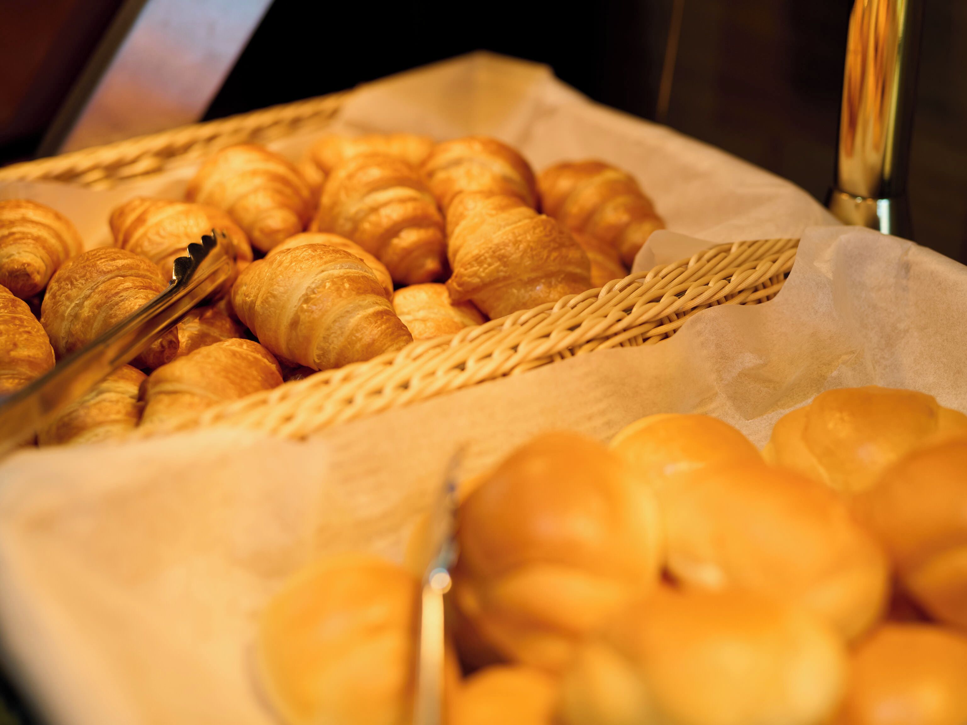 A display of assorted baked goods, including croissants and other pastries, arranged on a tray with a woven basket and a pair of tongs.