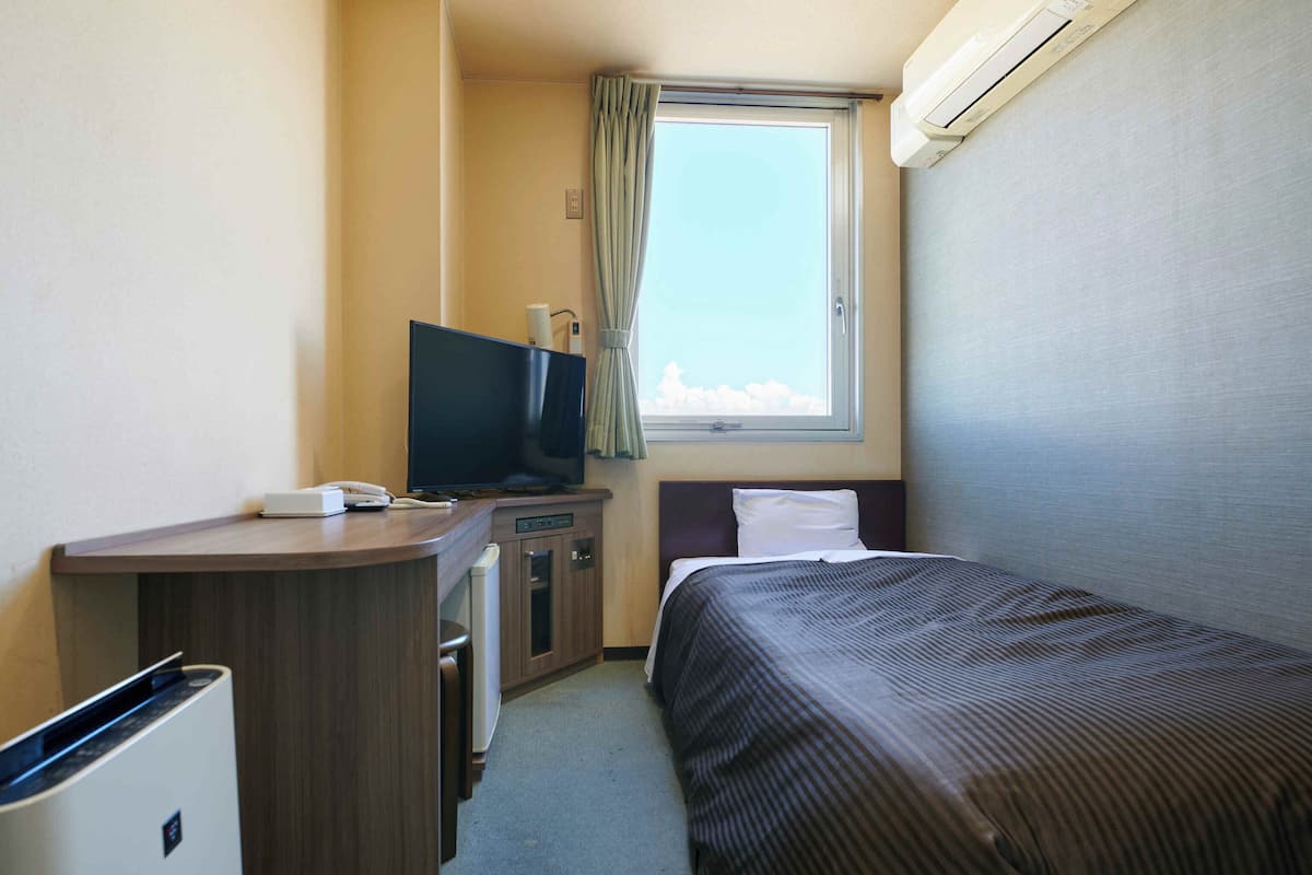 A compact hotel room with a bed, a desk with a television, a small kitchenette, and a window with curtains.
