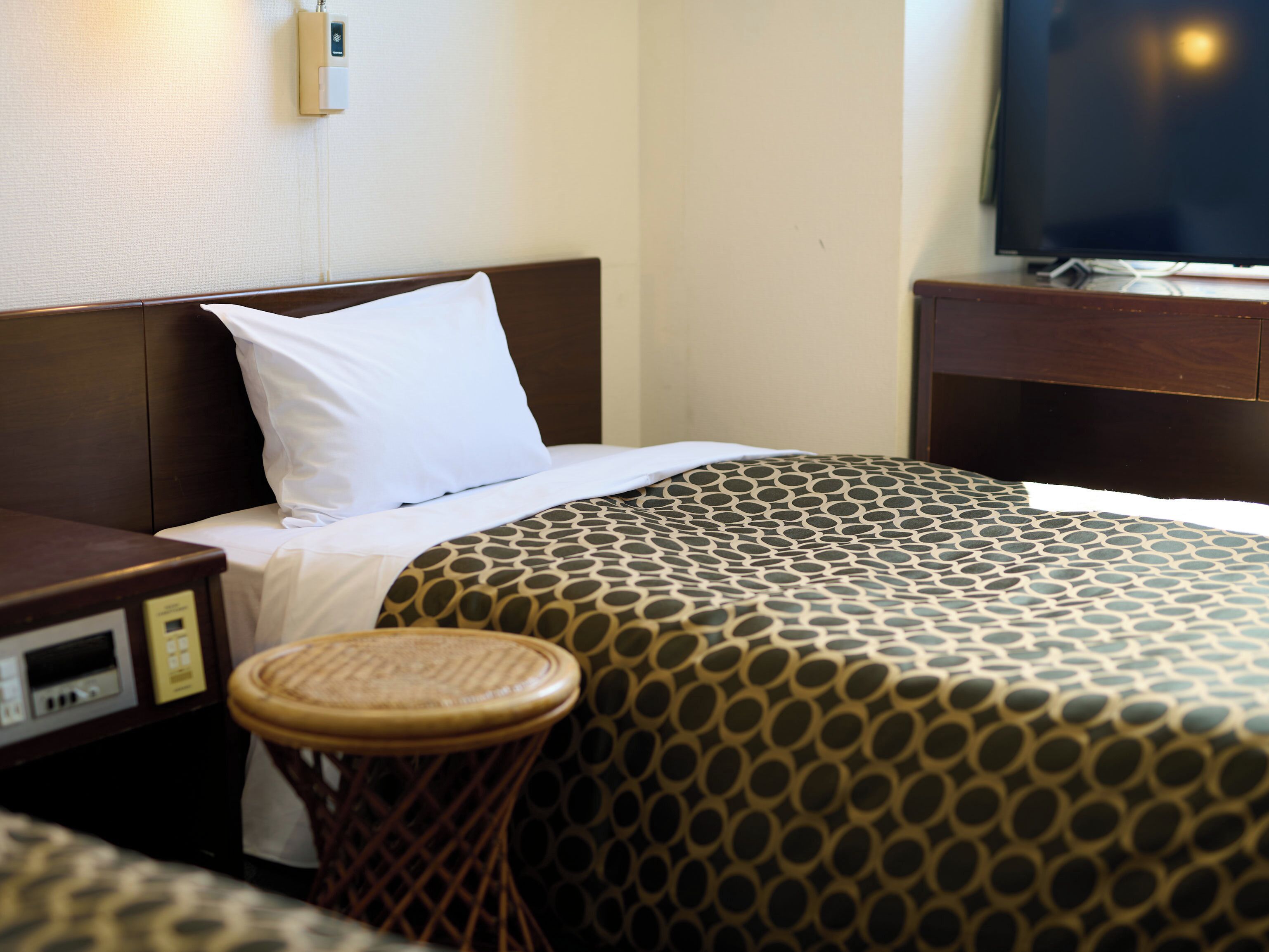 A hotel room with a bed, a bedside table, a television, and a small stool.