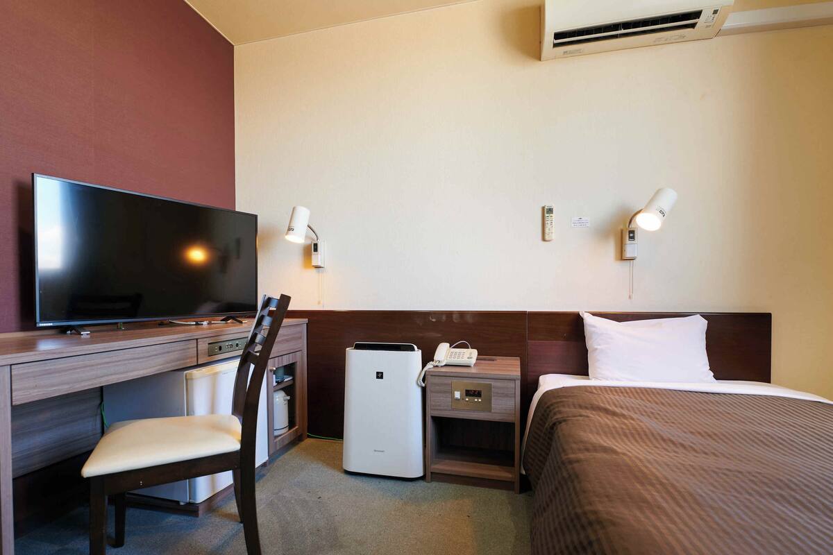 A hotel room with a flat-screen TV, a desk, a chair, a bed, bedside tables, lamps, and an air conditioner.