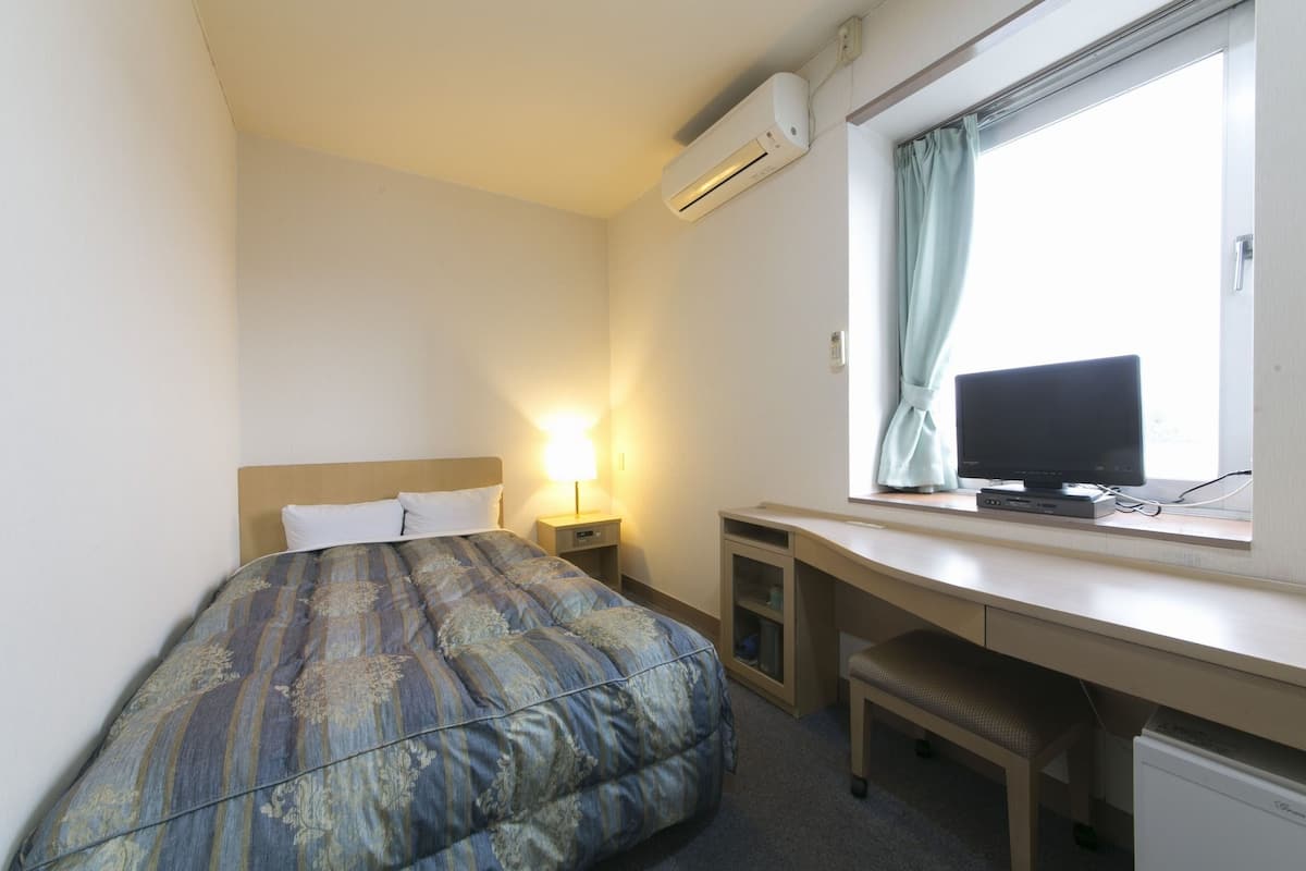 Ein Hotelzimmer mit einem Bett, einem Schreibtisch mit Computer, einem Fenster mit Vorhängen und einer Klimaanlage.