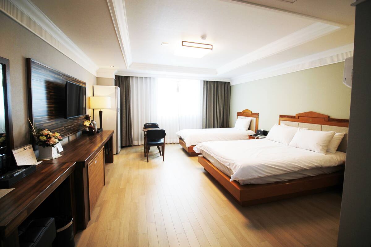 Quarto de hotel com escrivaninha de madeira, televisão de tela plana, duas camas, uma cadeira e janela com cortinas.