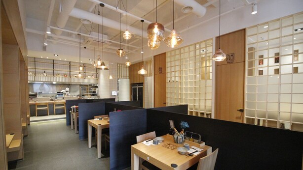 Un espace de restauration moderne avec des tables en bois et des cloisons noires, agrémenté de luminaires suspendus et d’une cuisine ouverte visible au fond.