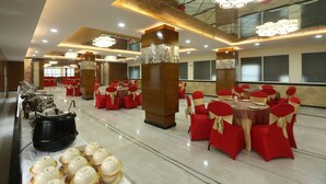 Una sala da banchetti con tavoli rotondi, sedie rosse e dorate, e un'area buffet con pentole e ciotole.