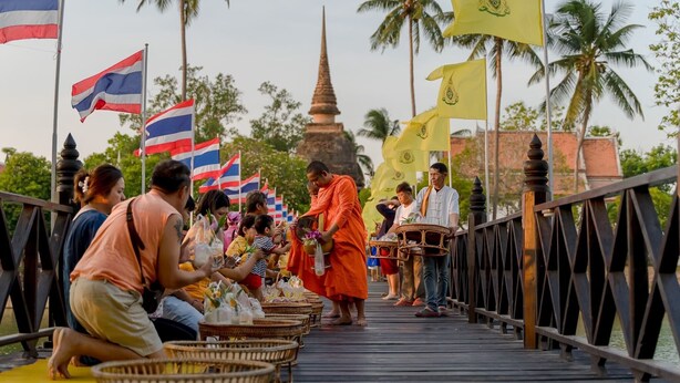 A wooden bridge with people and flags, surrounded by palm trees and a temple in the background. - Sukhothai Garden (Sukhothai)