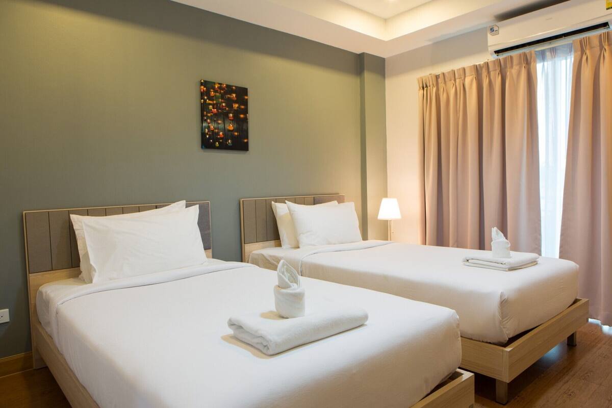 A hotel room with two beds, each with white linens and a folded towel on the bed. There is a wall-mounted air conditioner above the beds and a window with beige curtains.