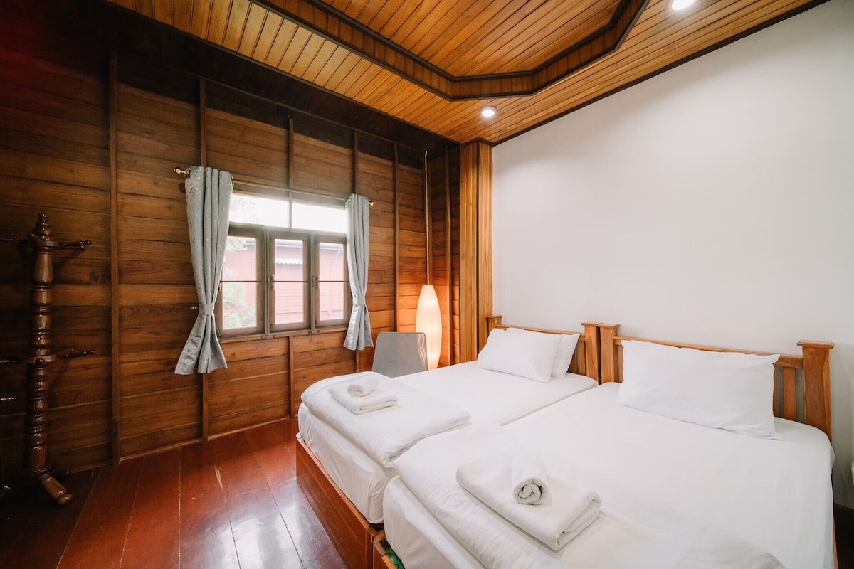 A room with wooden walls and ceiling, two beds with white linens, a wooden wardrobe, and a window with curtains.