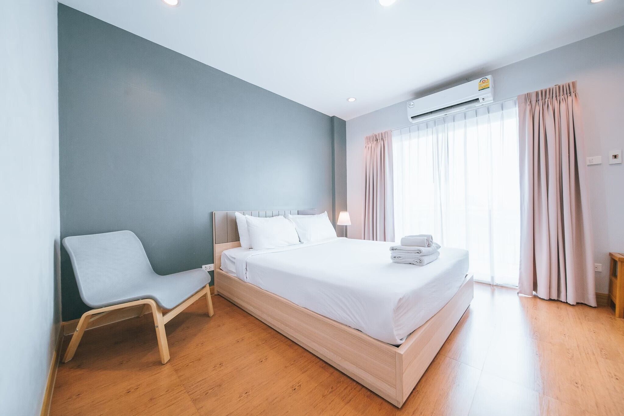 Moderni hotellihuone, jossa on yksi sänky, harmaa tuoli, puulattia ja suuri ikkuna, jossa on verhot.