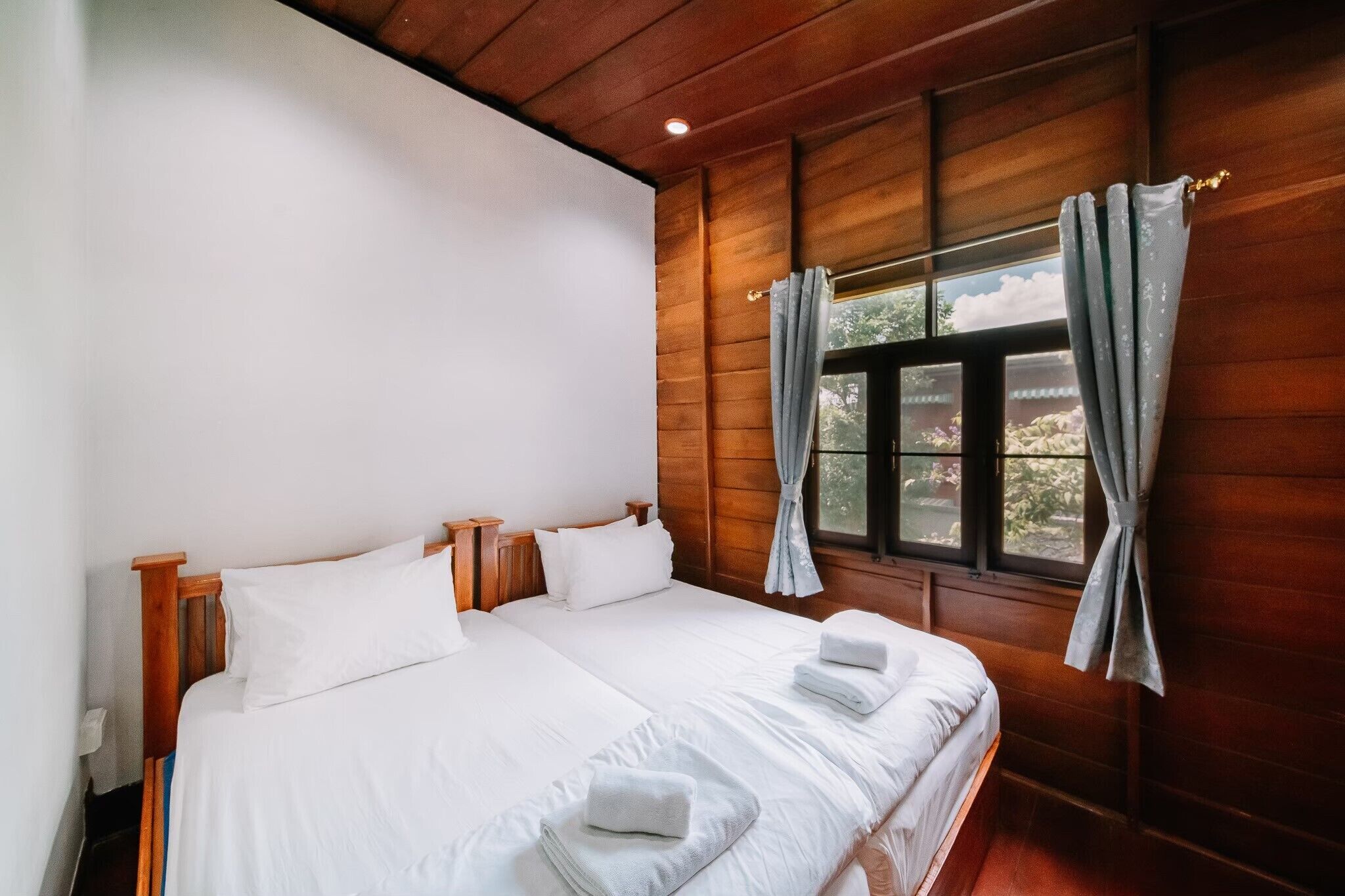 Cama doble con sábanas blancas y dos almohadas, cabecera de madera y ventana con cortinas.