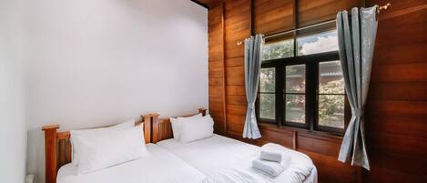Cama doble con sábanas blancas y dos almohadas, cabecera de madera y ventana con cortinas.