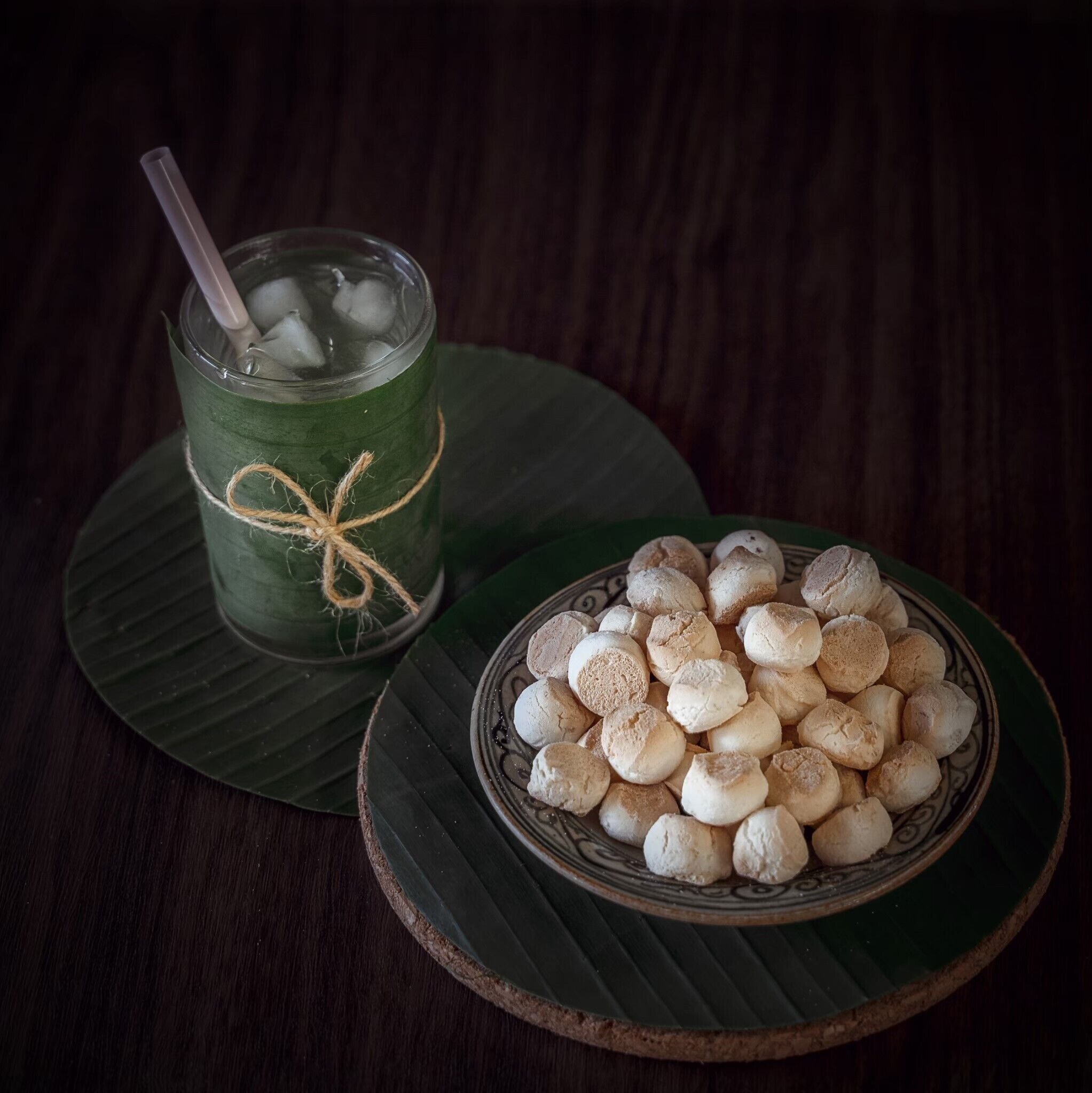 Un verre avec une paille et des glaçons, attaché avec une ficelle, posé sur une feuille verte. À côté, on aperçoit un bol rempli de biscuits ronds et blancs, disposés sur une assiette décorative.